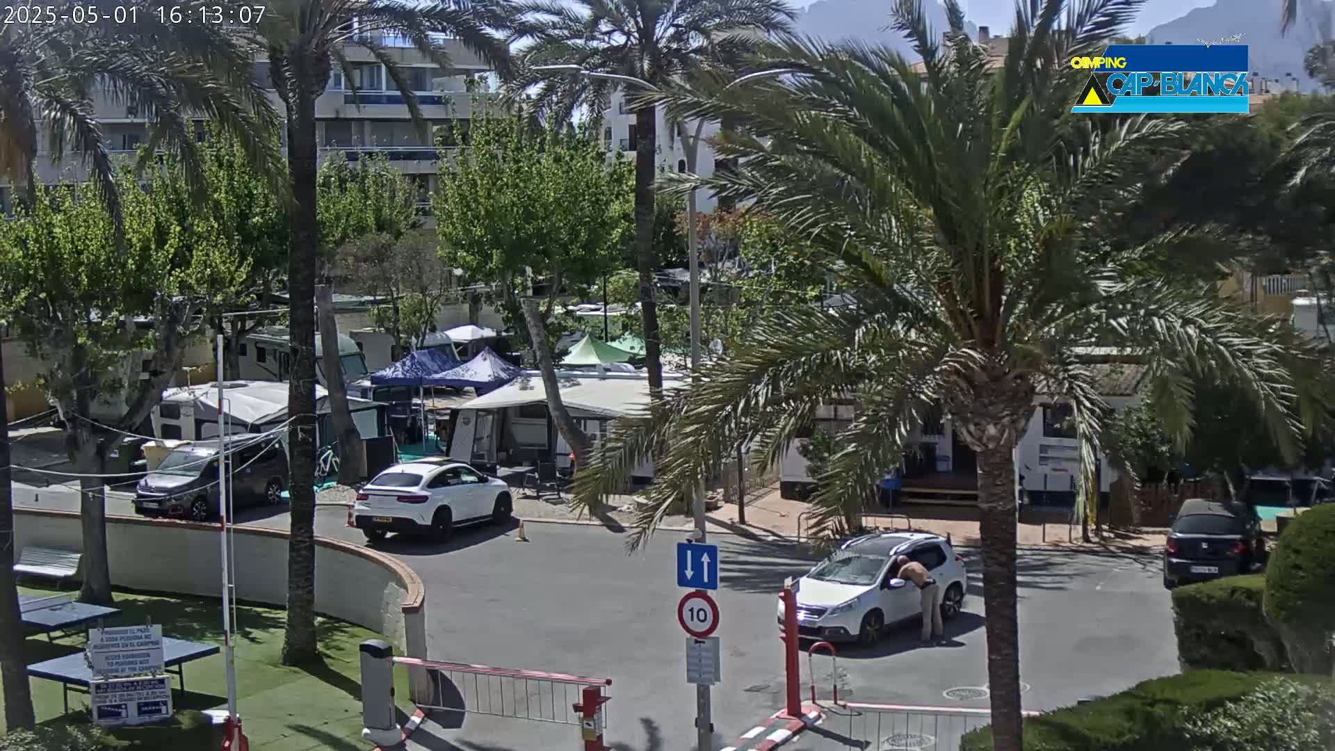 A sunny outdoor scene shows a parking lot with several cars and RVs amongst palm trees and buildings, near a gated entrance.