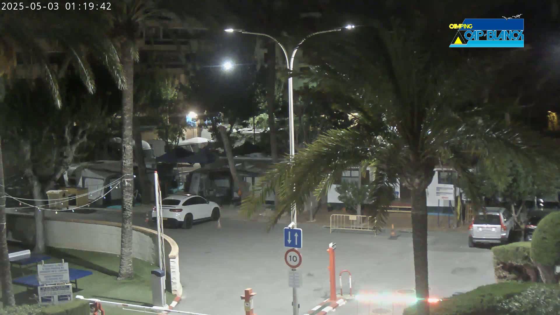 A nighttime view of a parking lot and roadway in a campground, surrounded by palm trees and buildings, with cars parked and a few people visible.