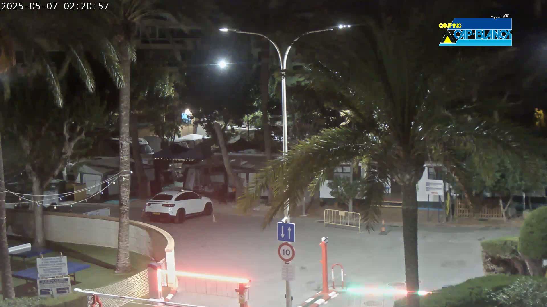 A nighttime view of a parking lot surrounded by palm trees and buildings, with a white car parked near a gate.