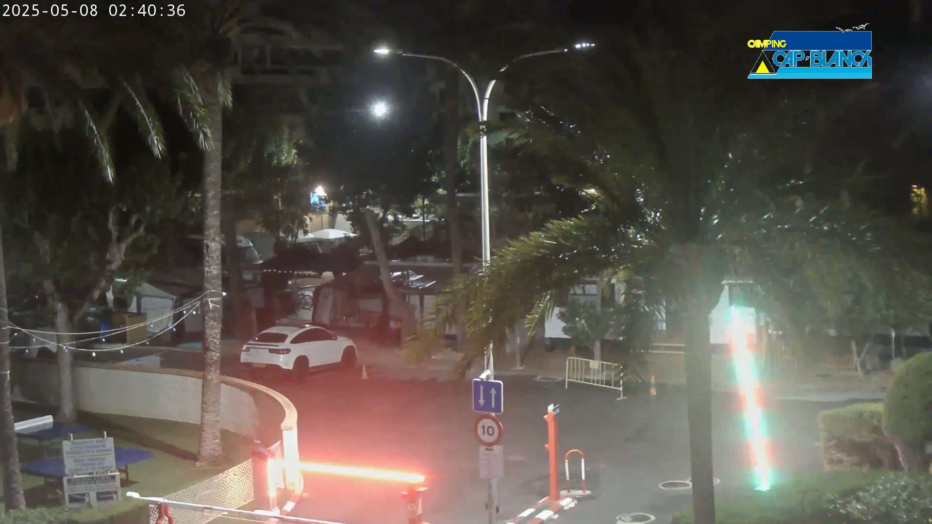 A nighttime scene shows a white SUV parked near a roadway with palm trees, low light levels, and a barrier with a red light.