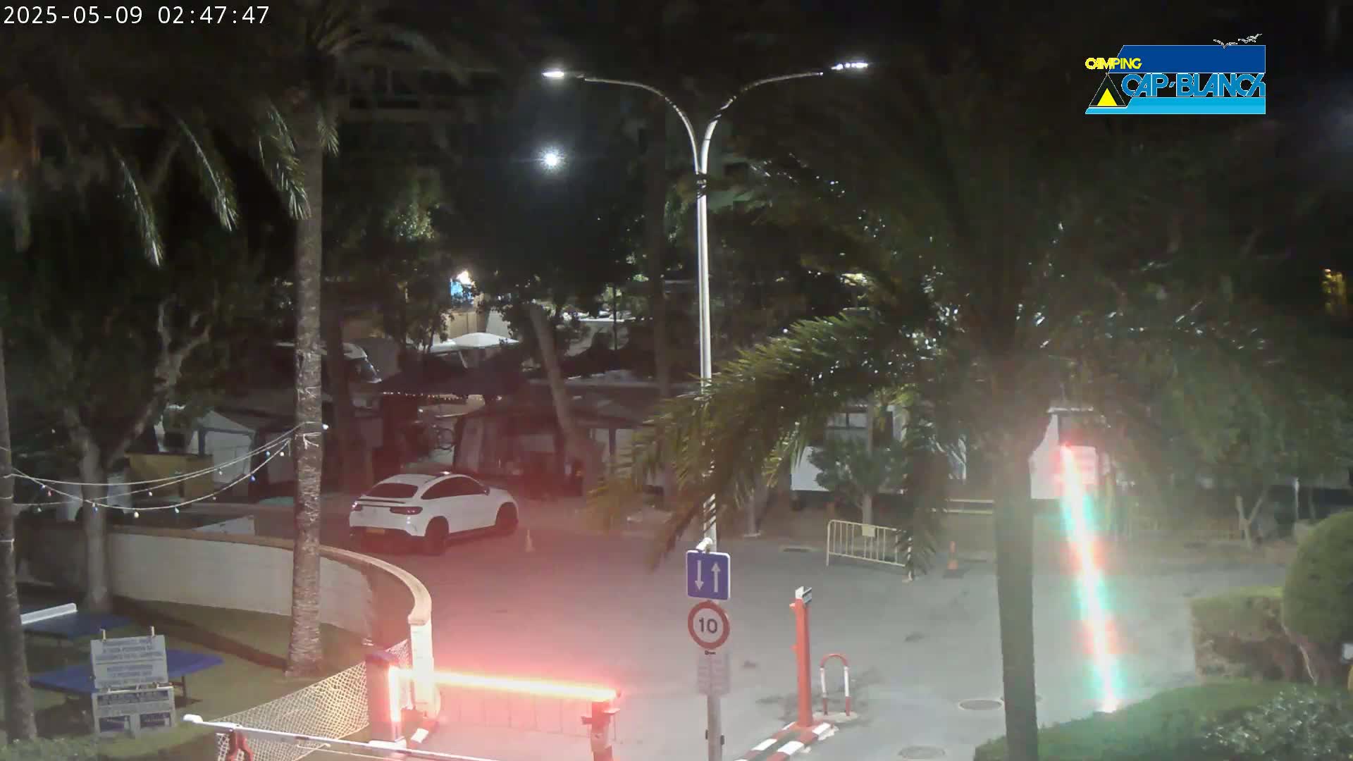 A nighttime view of a parking lot and surrounding area, illuminated by streetlights, showing palm trees, small buildings or trailers, and a white SUV.