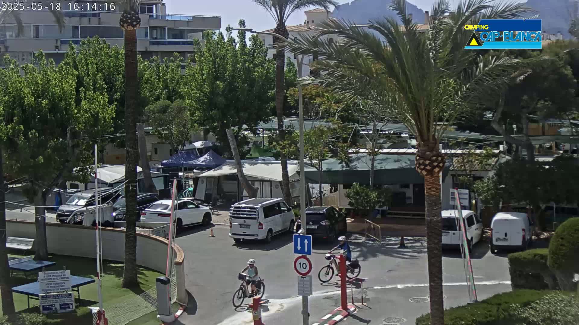 A sunny outdoor scene shows a campground with cars, campervans, and two people riding bicycles in a paved area near buildings and palm trees.