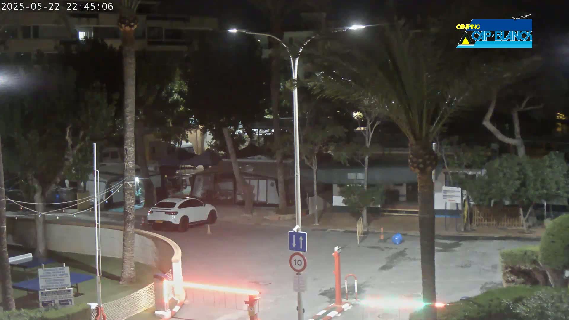 A nighttime view of a parking lot with a white car, palm trees, and buildings under artificial light.
