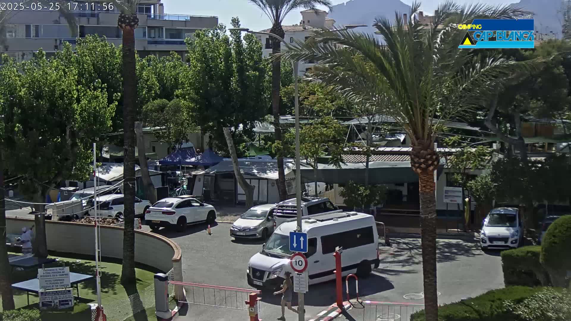 A sunny outdoor scene shows a parking lot with several cars and a white van, situated near buildings, palm trees, and what appears to be a campground with tents and small structures.