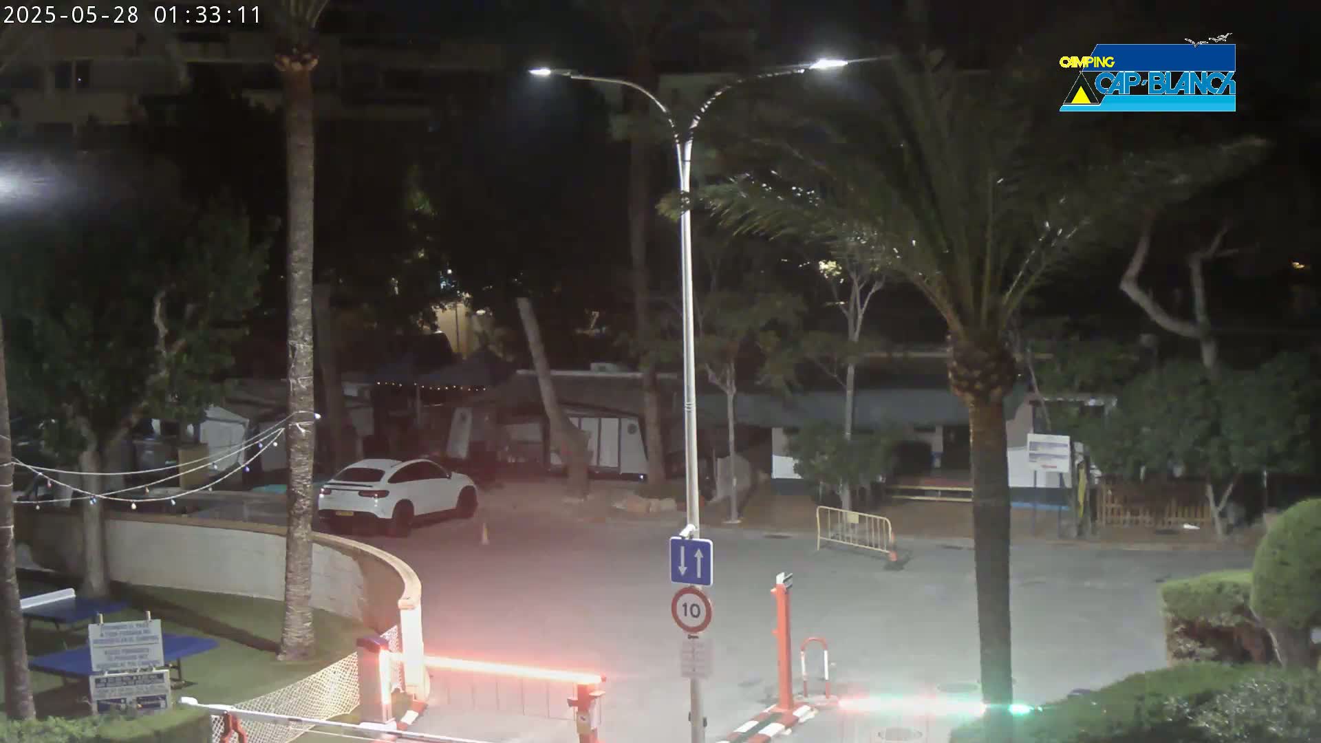 A nighttime scene shows a white SUV parked near a street light in a palm tree-lined area with a security gate and a speed limit sign.