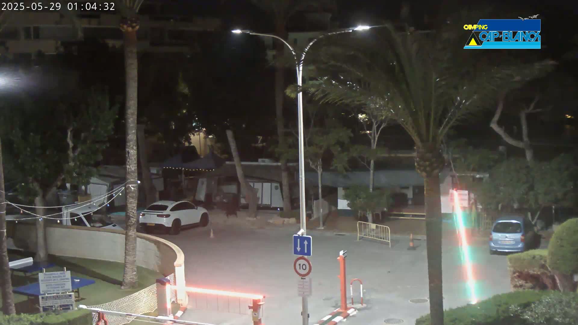 A nighttime scene of a parking lot with palm trees, streetlights, and a few parked cars, including a white SUV.