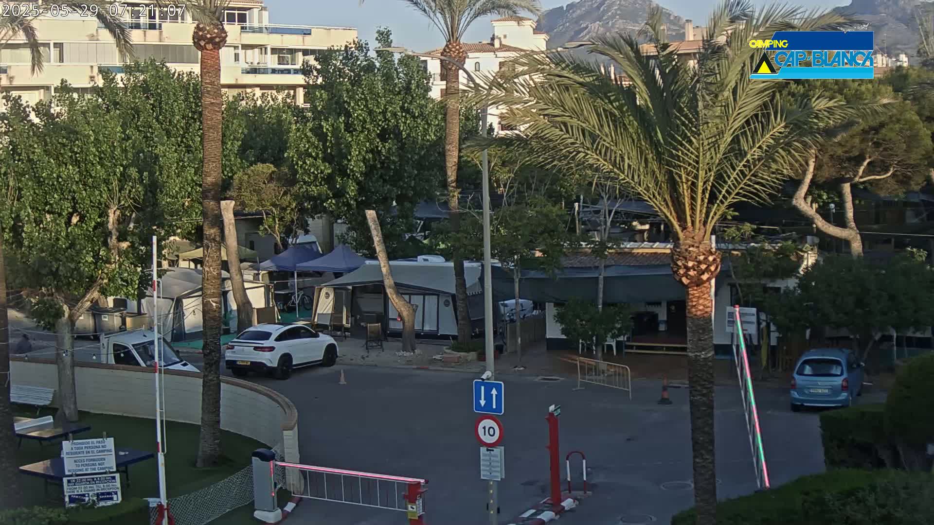 A sunny outdoor scene shows a roadway entering a campground with several RVs, tents, and cars, surrounded by palm trees and buildings.