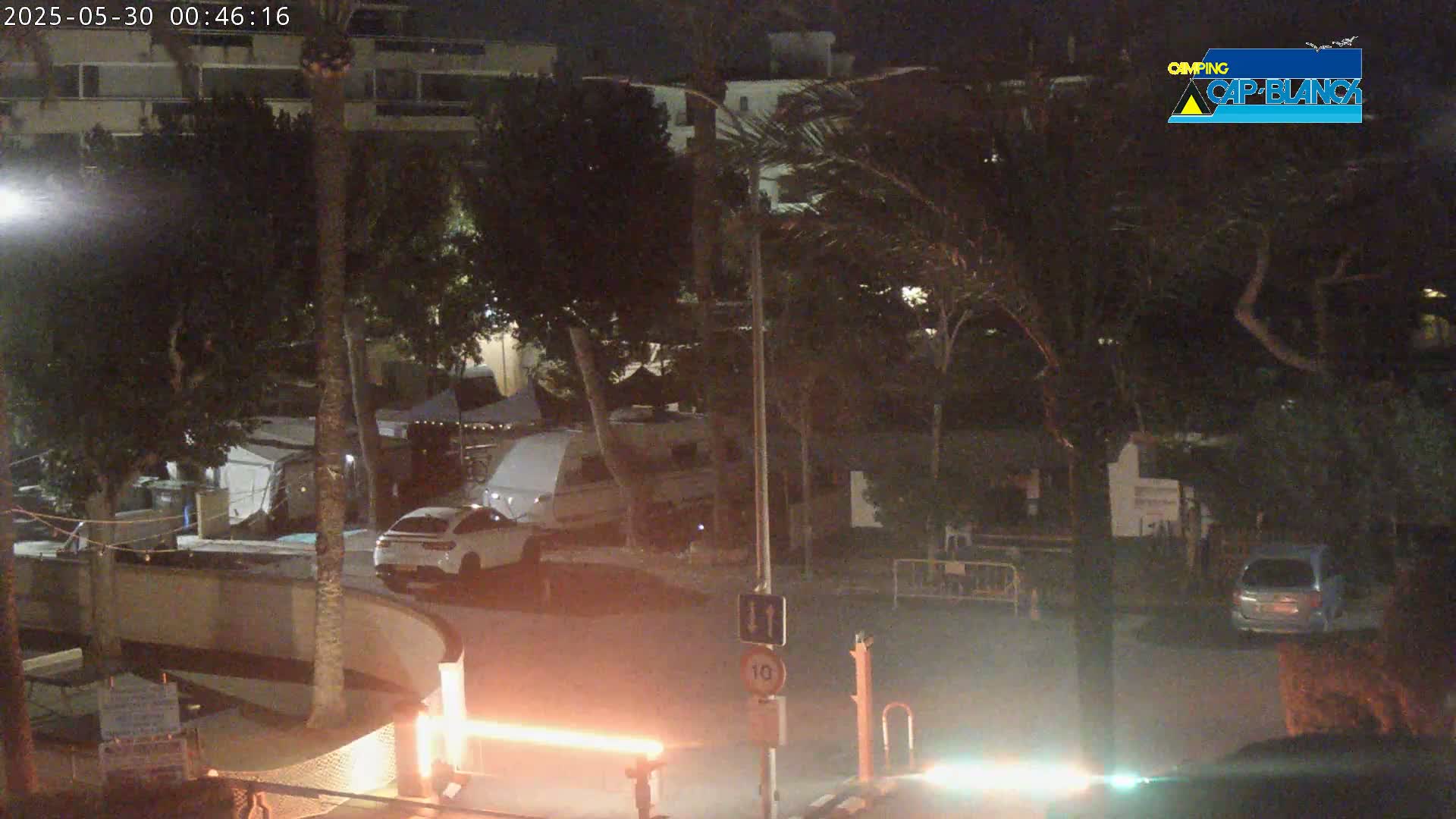 A nighttime outdoor scene shows a parking lot with cars and an RV, surrounded by trees and a building, under windy conditions.