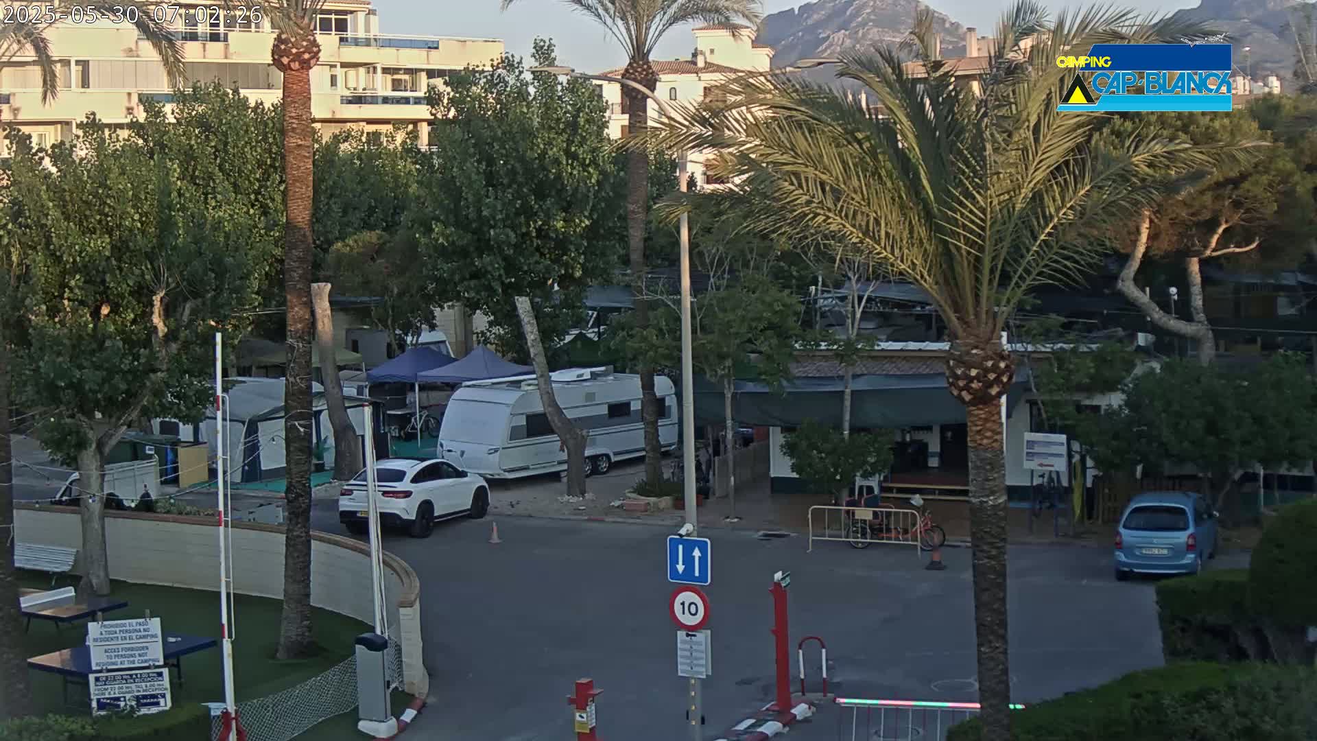 A campground with several caravans and cars, palm trees, and buildings under a partly cloudy sky.