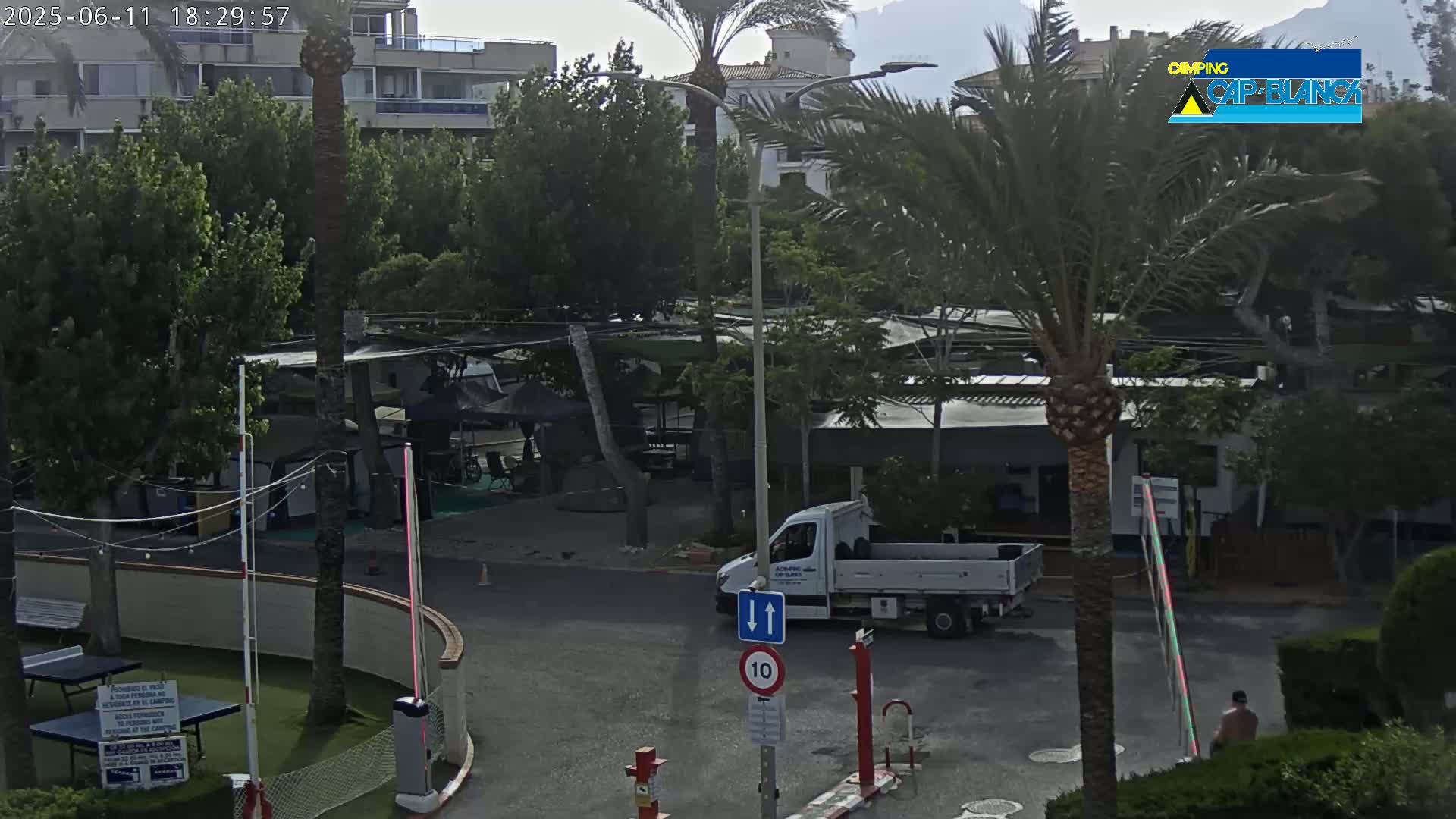 A mostly cloudy day reveals a parking lot and road near a campground with several buildings and palm trees, with a white truck parked near a speed limit sign.