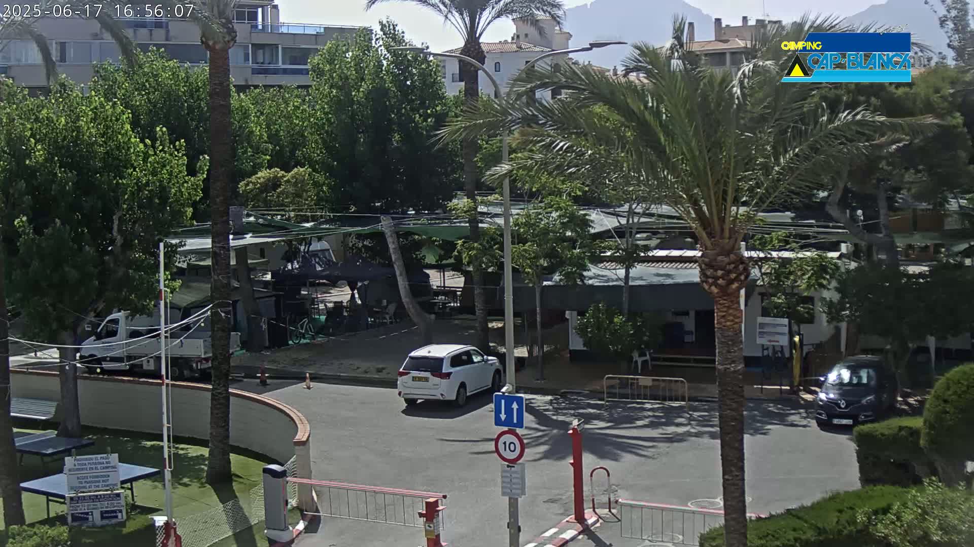 A sunny outdoor scene shows a roadway with several vehicles, including a white SUV, near a campground with palm trees and buildings under a mostly clear sky.