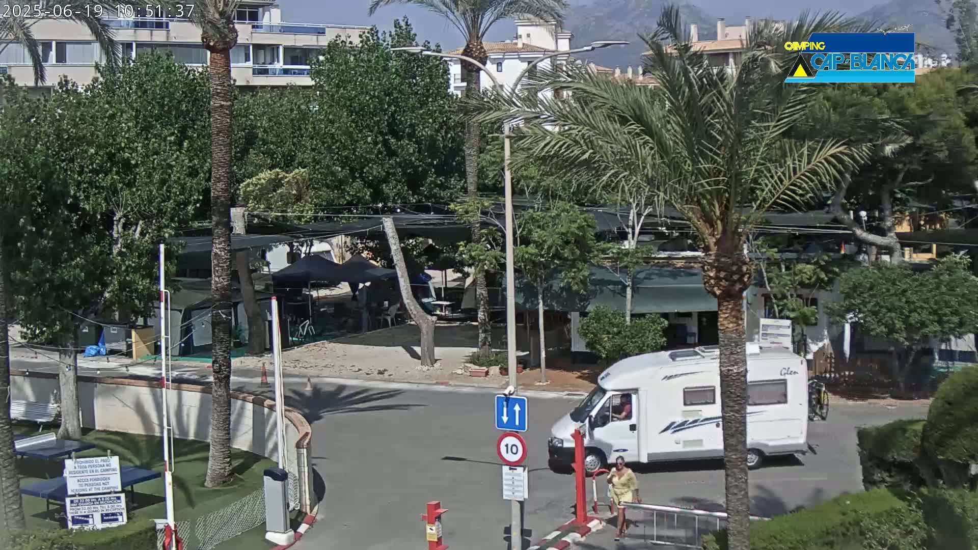A sunny scene shows a roadway intersection in a campground with palm trees, buildings, RVs, and a few people.