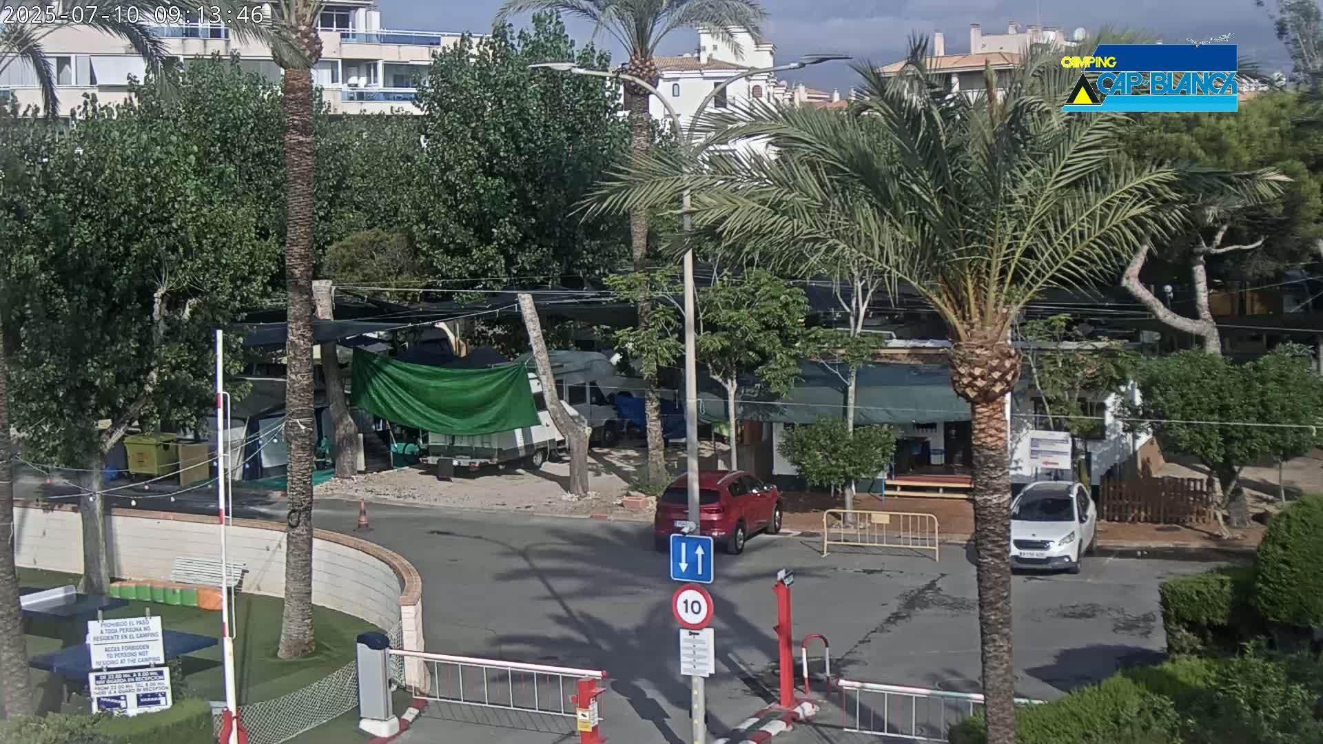 A sunny outdoor scene shows a paved area with cars parked near buildings and lush greenery, including palm trees, in a campground setting.