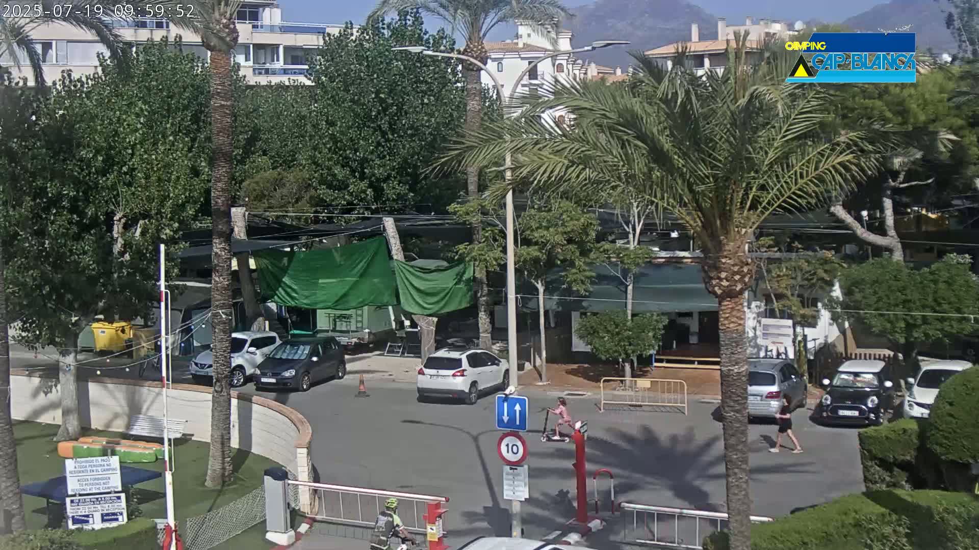 A sunny outdoor scene shows a street with parked cars, pedestrians, and cyclists amongst palm trees and buildings.