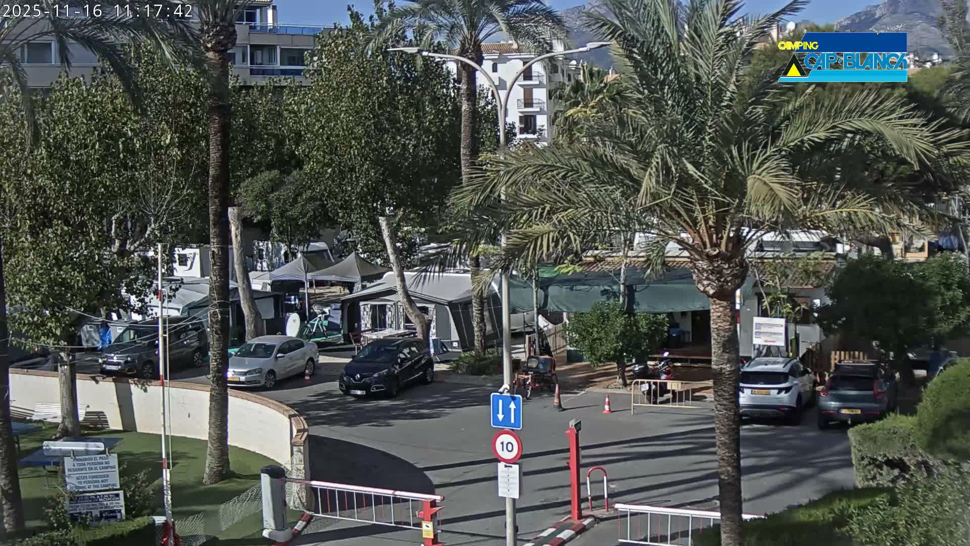 Under a clear blue sky and bright sunshine, an outdoor scene captures the entrance to what appears to be a campground or resort, with several cars, palm trees, barrier gates, and distant mountains.