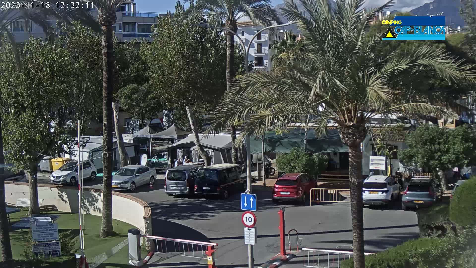 Under a clear blue sky and bright sunshine, an outdoor scene captures the entrance to what appears to be a campground or resort, with several cars, palm trees, barrier gates, and distant mountains.