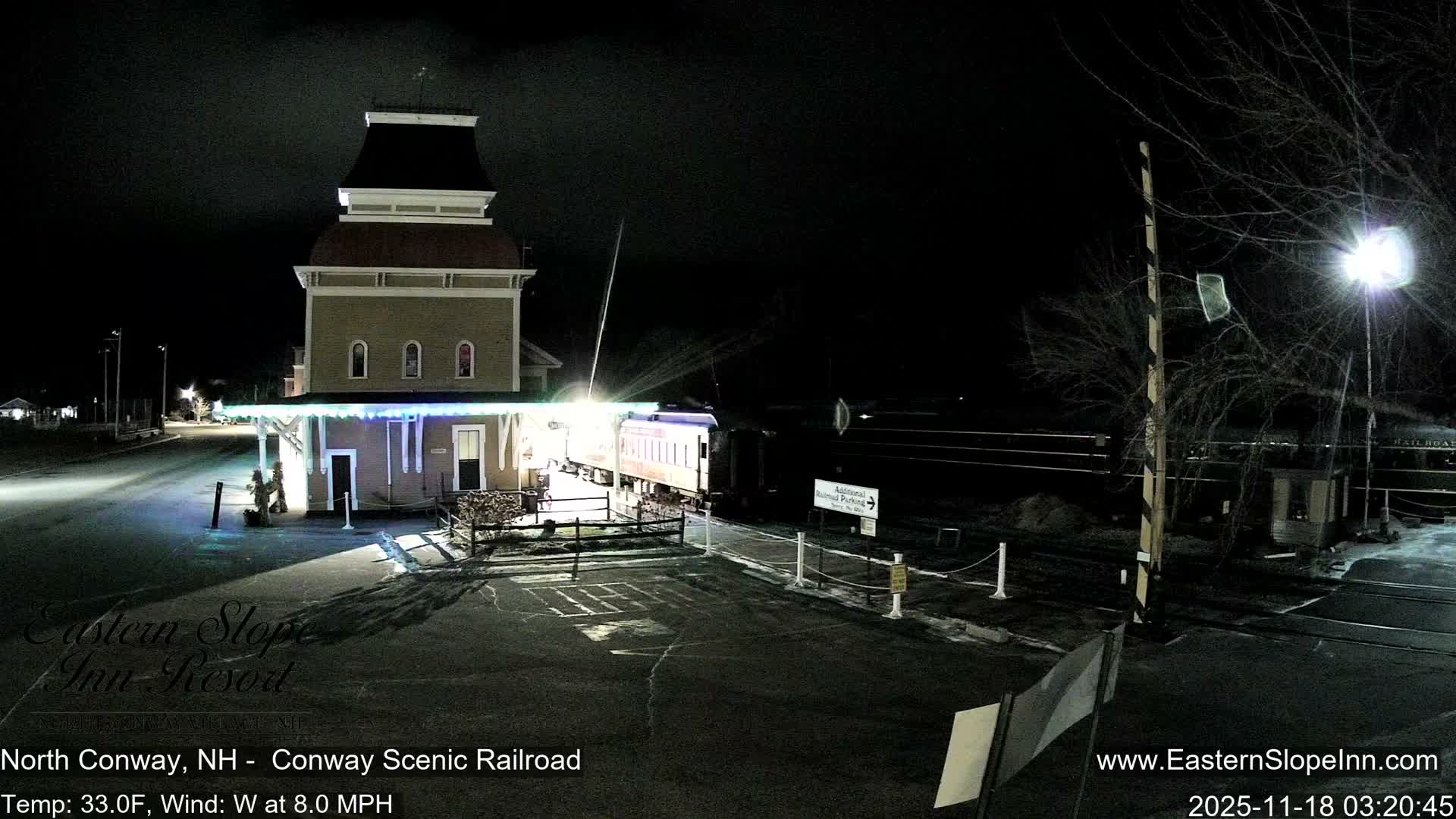A brightly illuminated, ornate train station stands beside a parked train on a clear, dark night, with a railroad crossing barrier and streetlights visible.