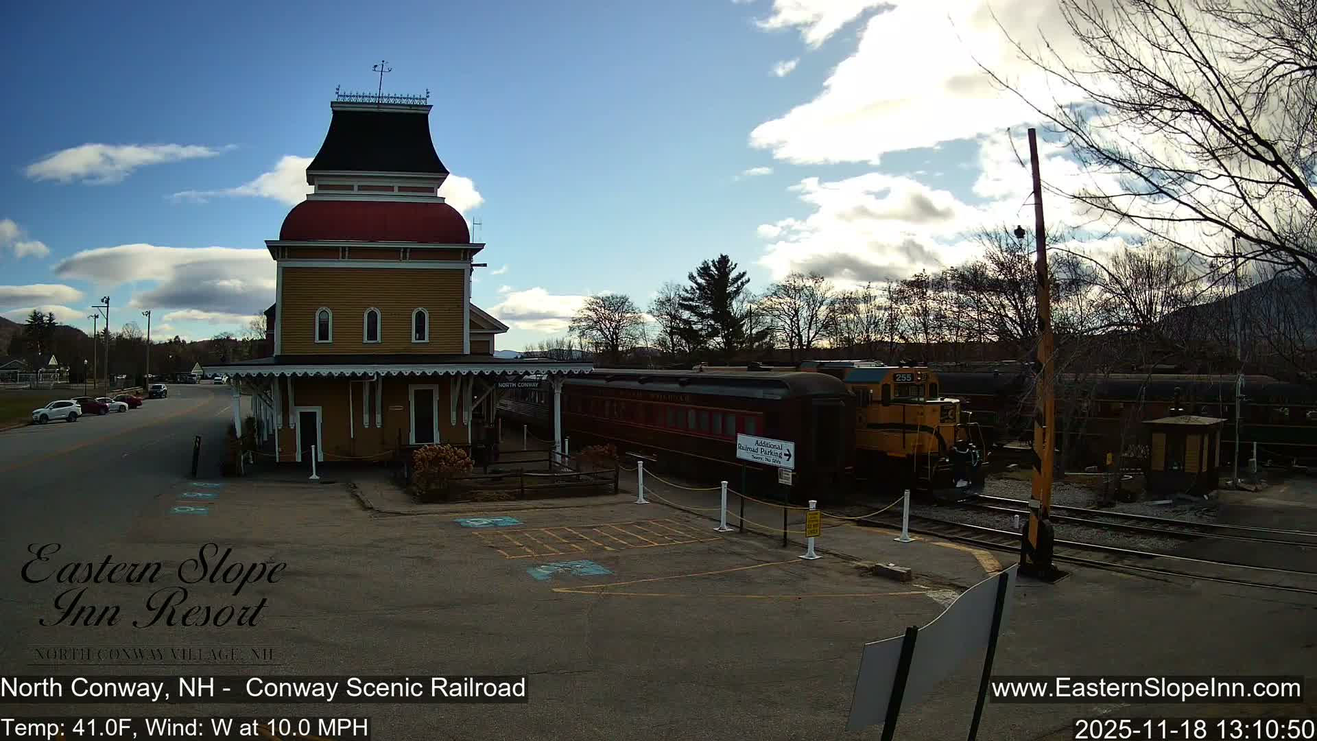 A brightly illuminated, ornate train station stands beside a parked train on a clear, dark night, with a railroad crossing barrier and streetlights visible.