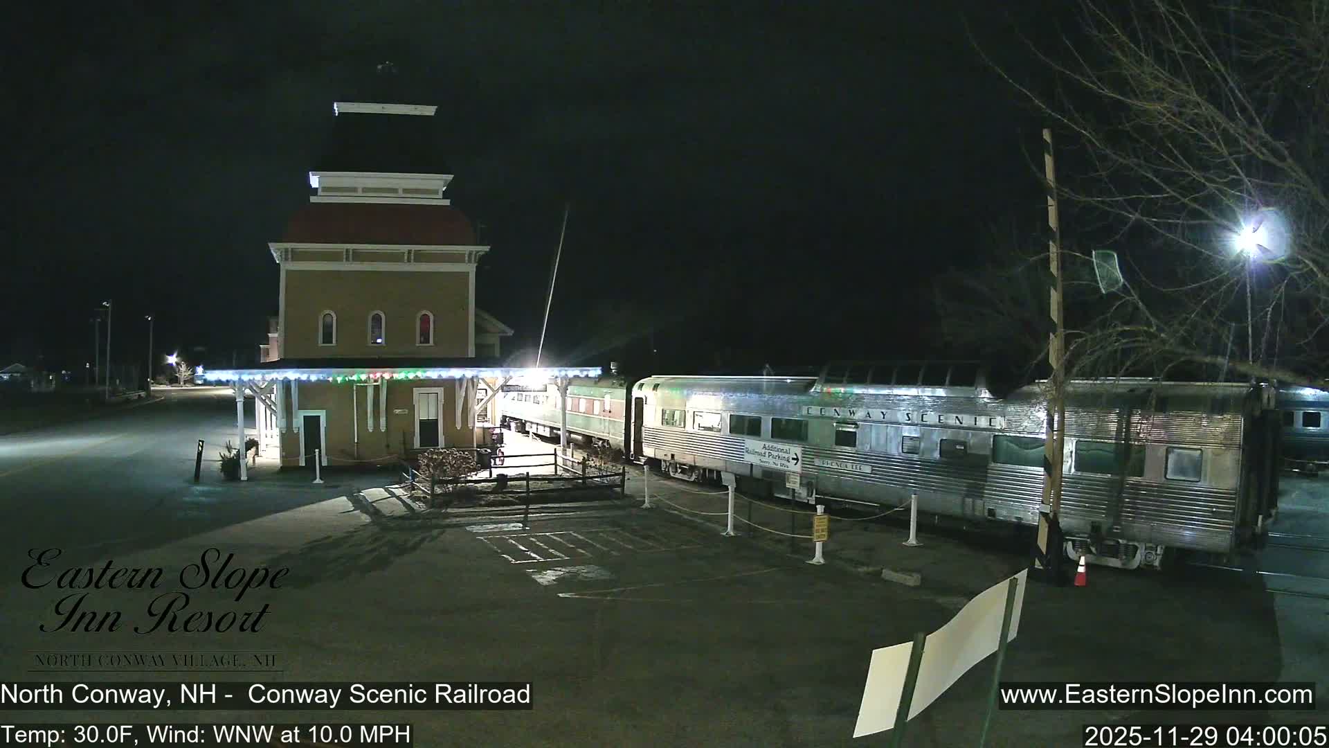 A multi-story train station building with a red roof stands beside a long silver and green passenger train parked on tracks, all illuminated under a clear night sky.