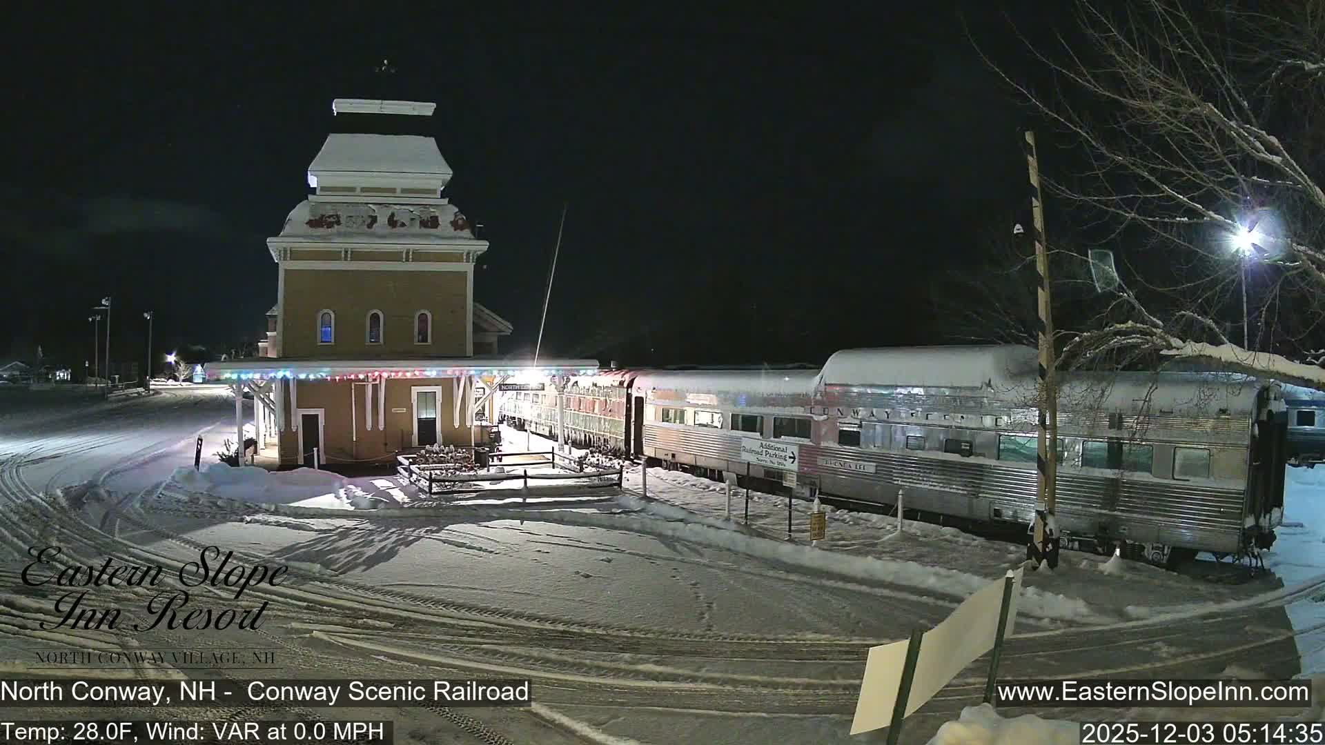 A snowy train station with a distinctive cupola stands next to a stationary passenger train under a dark, clear night sky, with vehicle tracks visible in the snow-covered foreground.