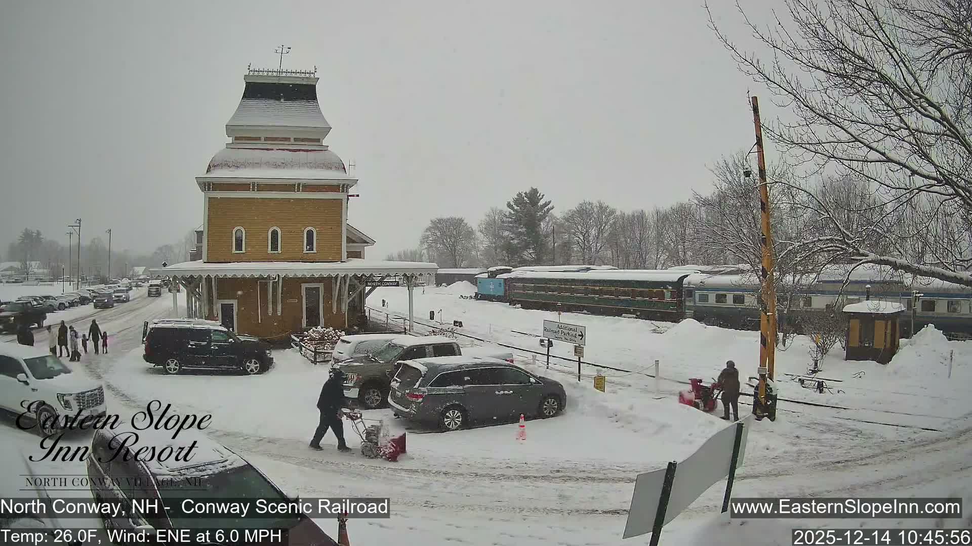 A snowy train station with a distinctive cupola stands next to a stationary passenger train under a dark, clear night sky, with vehicle tracks visible in the snow-covered foreground.