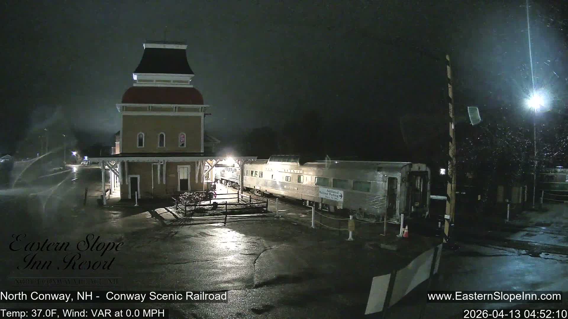 A snowy train station with a distinctive cupola stands next to a stationary passenger train under a dark, clear night sky, with vehicle tracks visible in the snow-covered foreground.