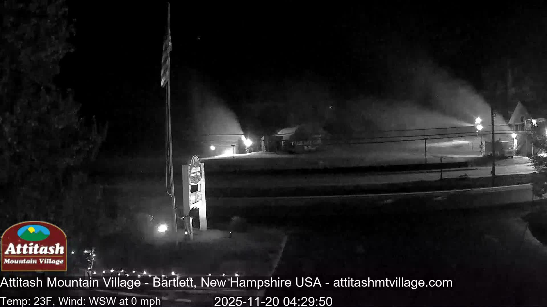 A nighttime view of a ski resort village shows multiple snow cannons actively spraying artificial snow, illuminated by their own lights and nearby street lamps under clear and very cold conditions.