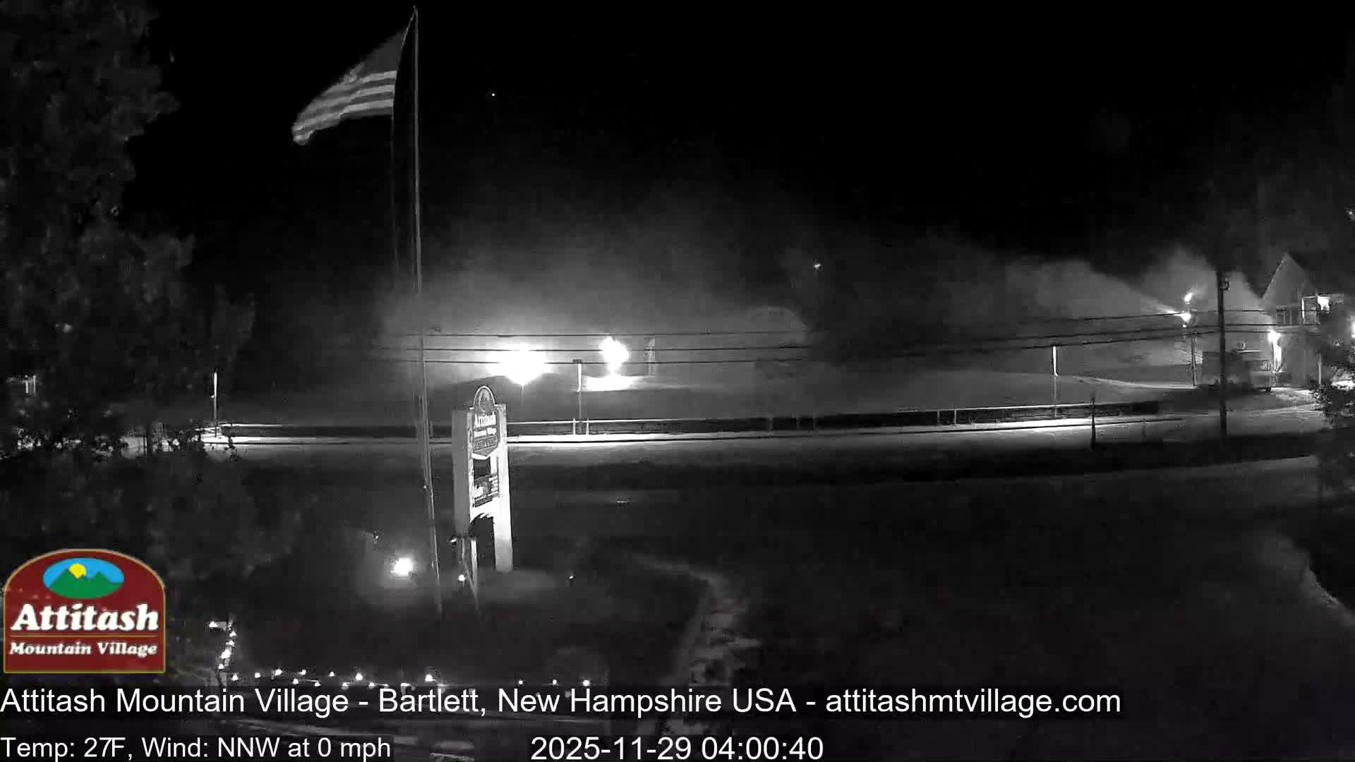 At night, under clear, cold conditions, an American flag waves above trees on the left, as bright lights illuminate a wide cloud of mist from active snowmaking operations blanketing a ski slope, with buildings visible in the distance.