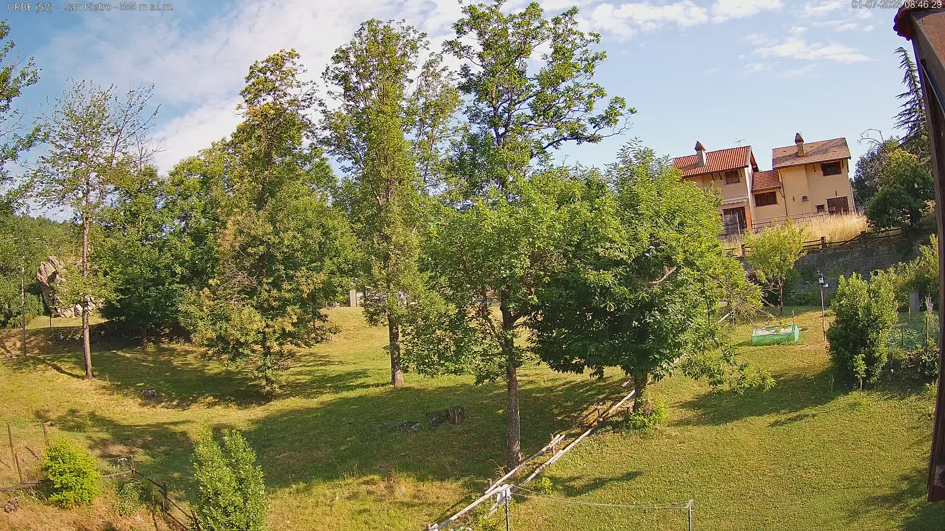A grassy yard with several deciduous trees partially obscures a view of a tan house in the distance under a partly cloudy sky.