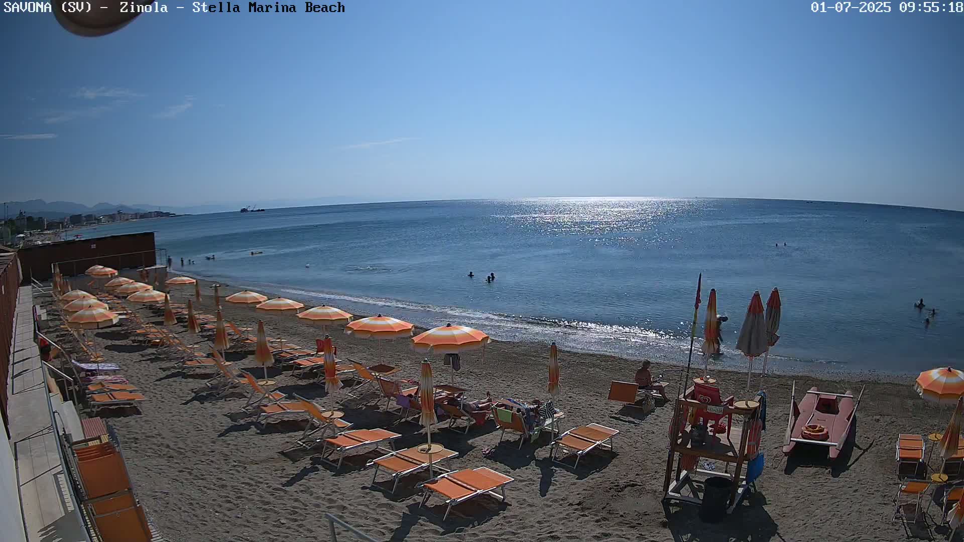A sandy beach with orange umbrellas and lounge chairs is shown, with calm blue water and a few people swimming in the sea on a sunny day.