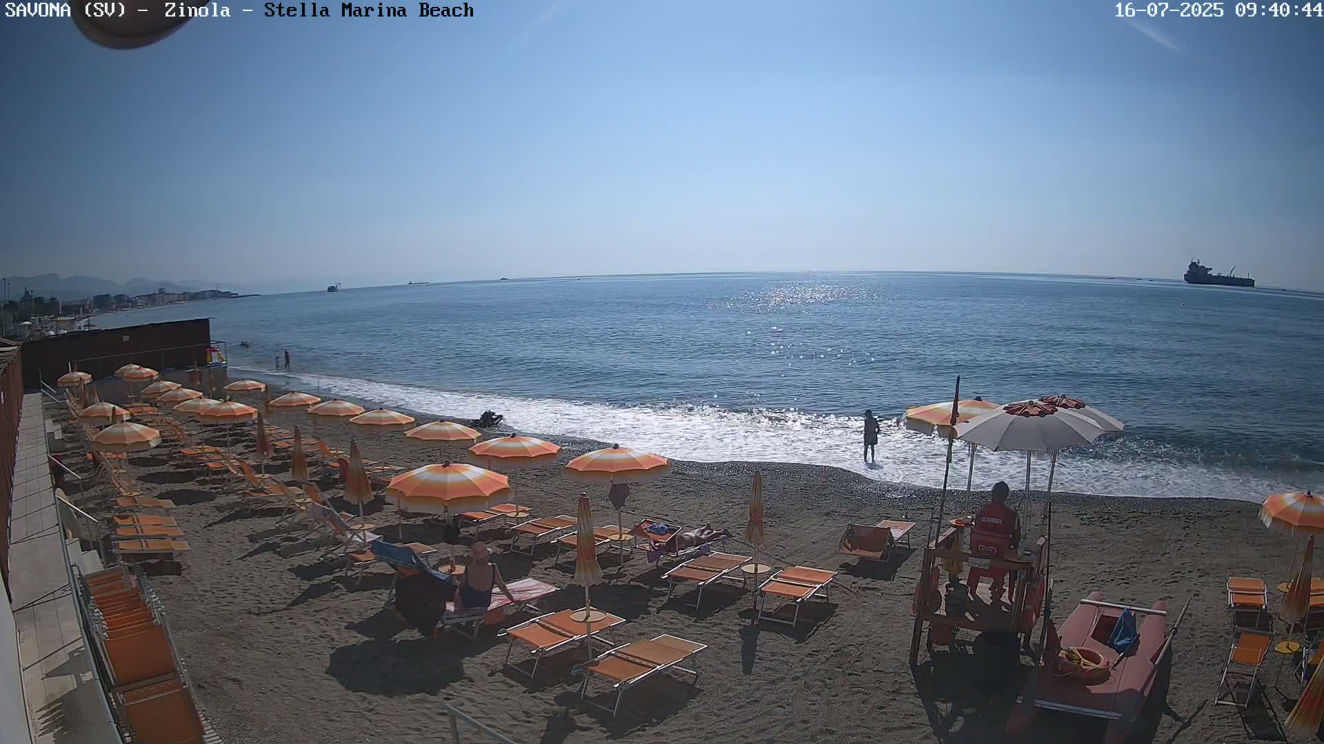 A sandy beach with orange umbrellas and lounge chairs is seen under a clear blue sky, with calm waves lapping the shore and a lifeguard station present.