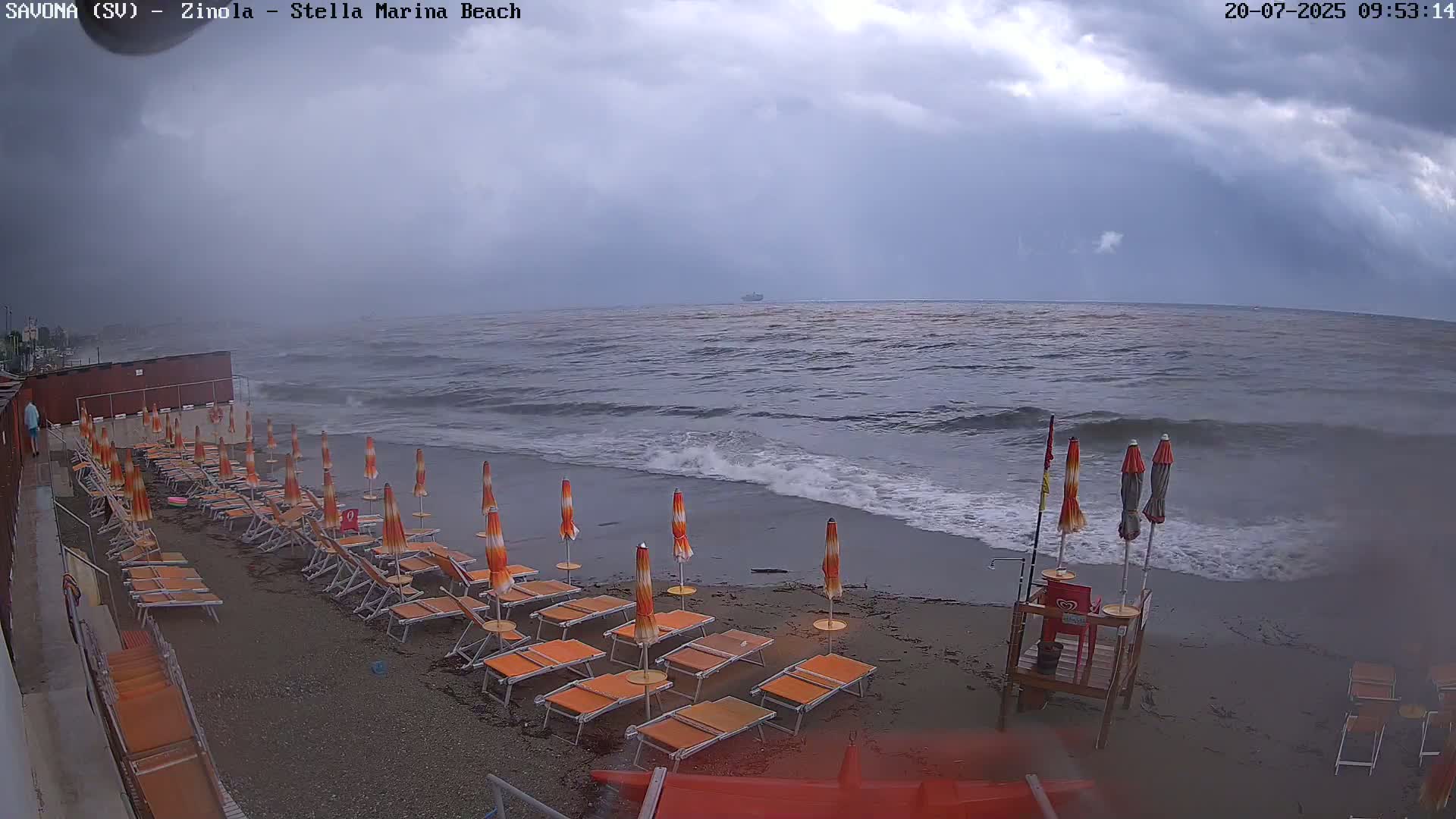 Rows of orange beach umbrellas and lounge chairs sit on a dark sand beach under a cloudy sky, with choppy waves rolling onto the shore.