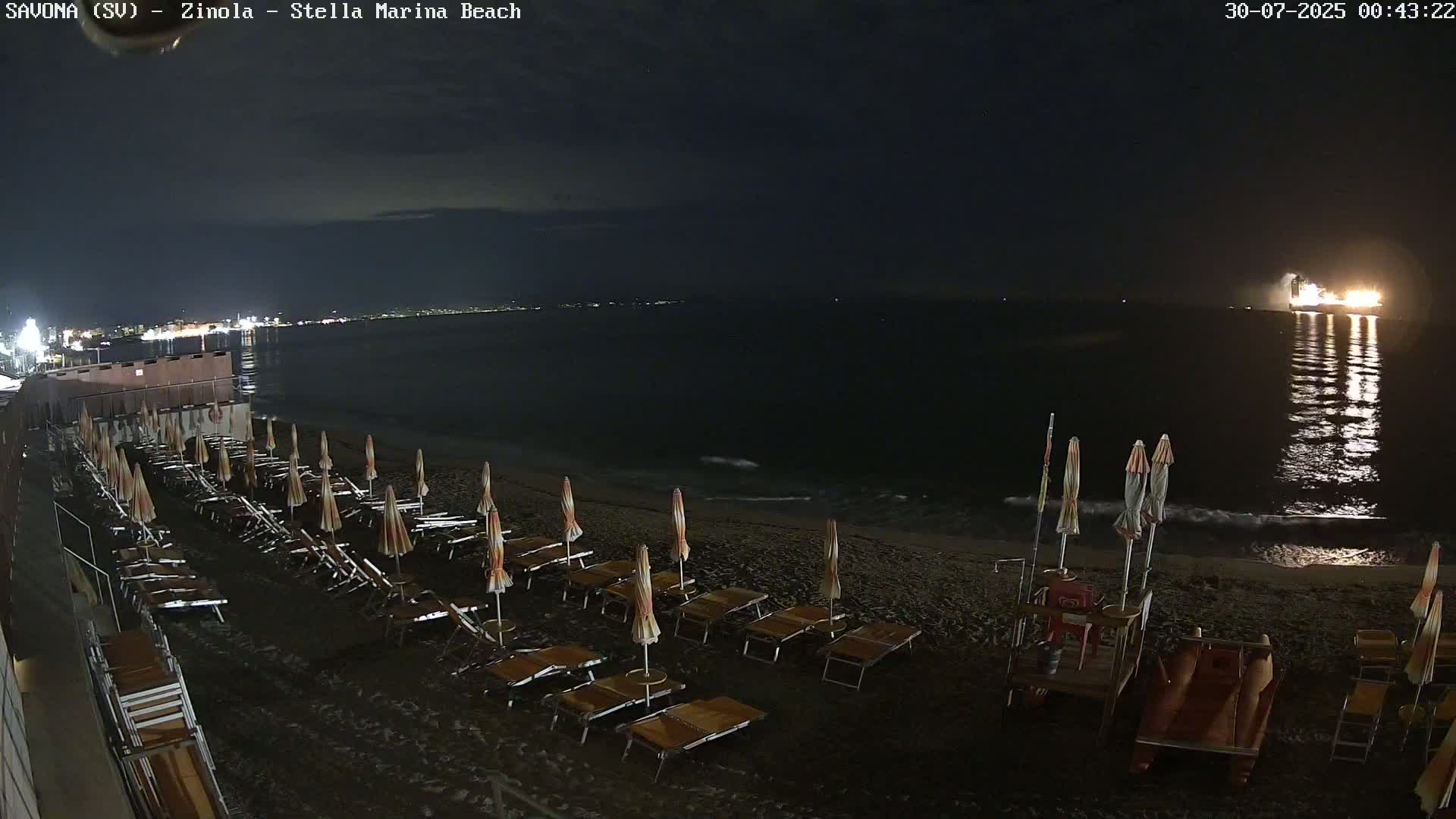 Rows of orange beach umbrellas and lounge chairs sit on a dark sand beach under a cloudy sky, with choppy waves rolling onto the shore.