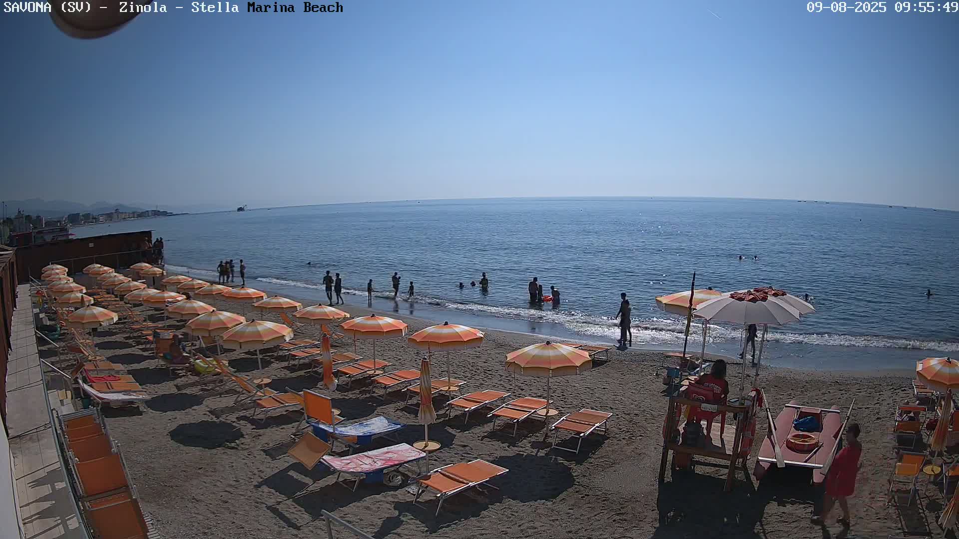A sandy beach with orange umbrellas and lounge chairs is partially occupied by people swimming and relaxing on a sunny day.