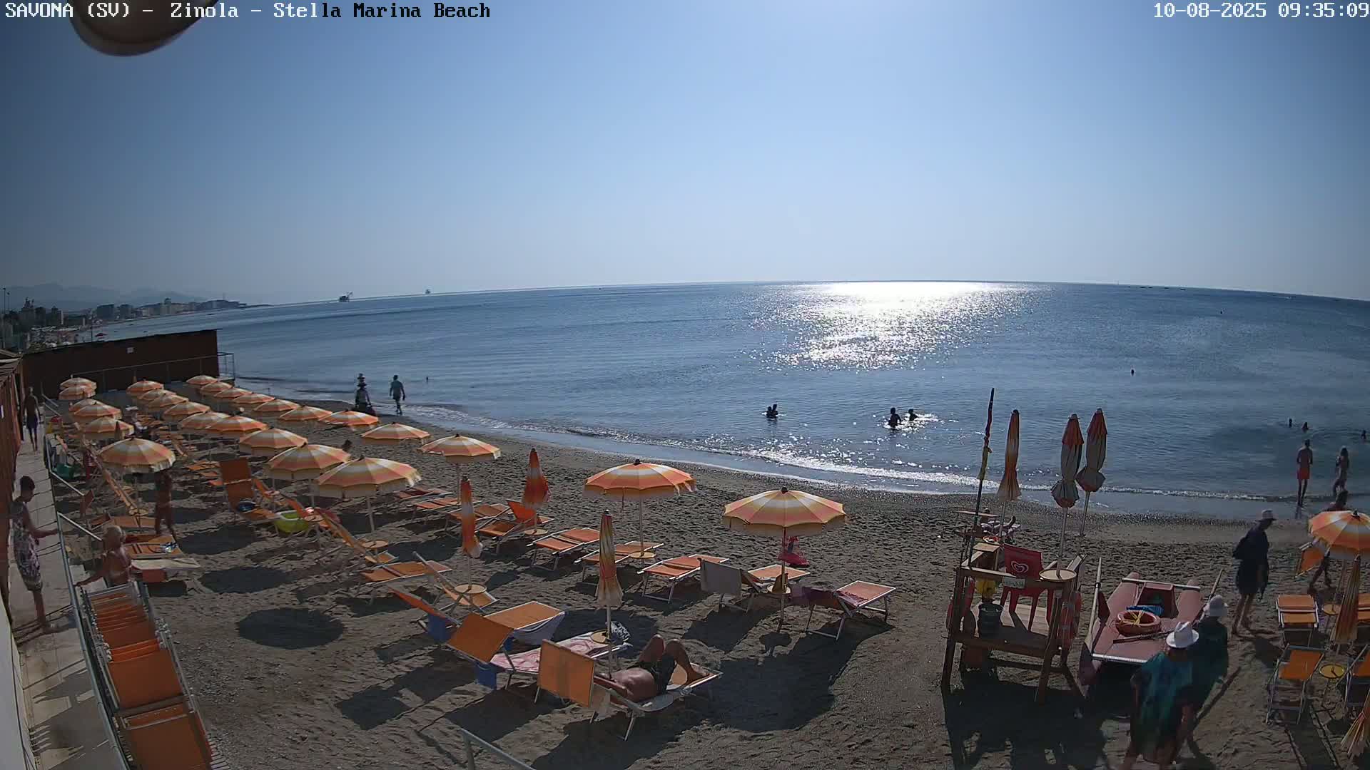 A sandy beach with numerous orange and white striped umbrellas and lounge chairs is shown, under a clear sunny sky, with several people swimming in the calm blue sea.