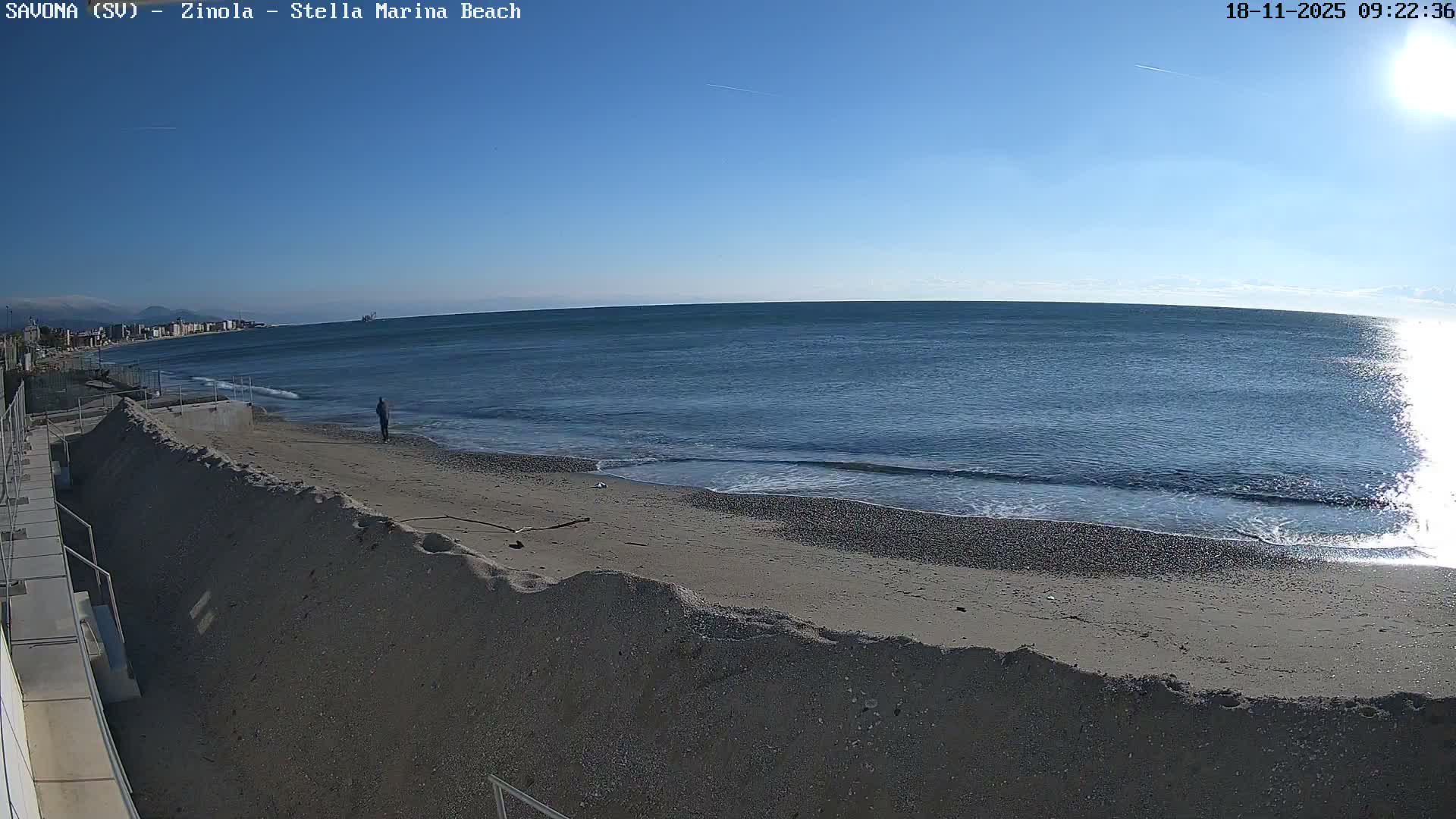 On a bright and sunny day, a lone person walks along a wide sandy and pebbled beach with gentle waves rolling in, while the sun gleams intensely on the ocean's surface and a distant coastal town lines the horizon under a clear blue sky.