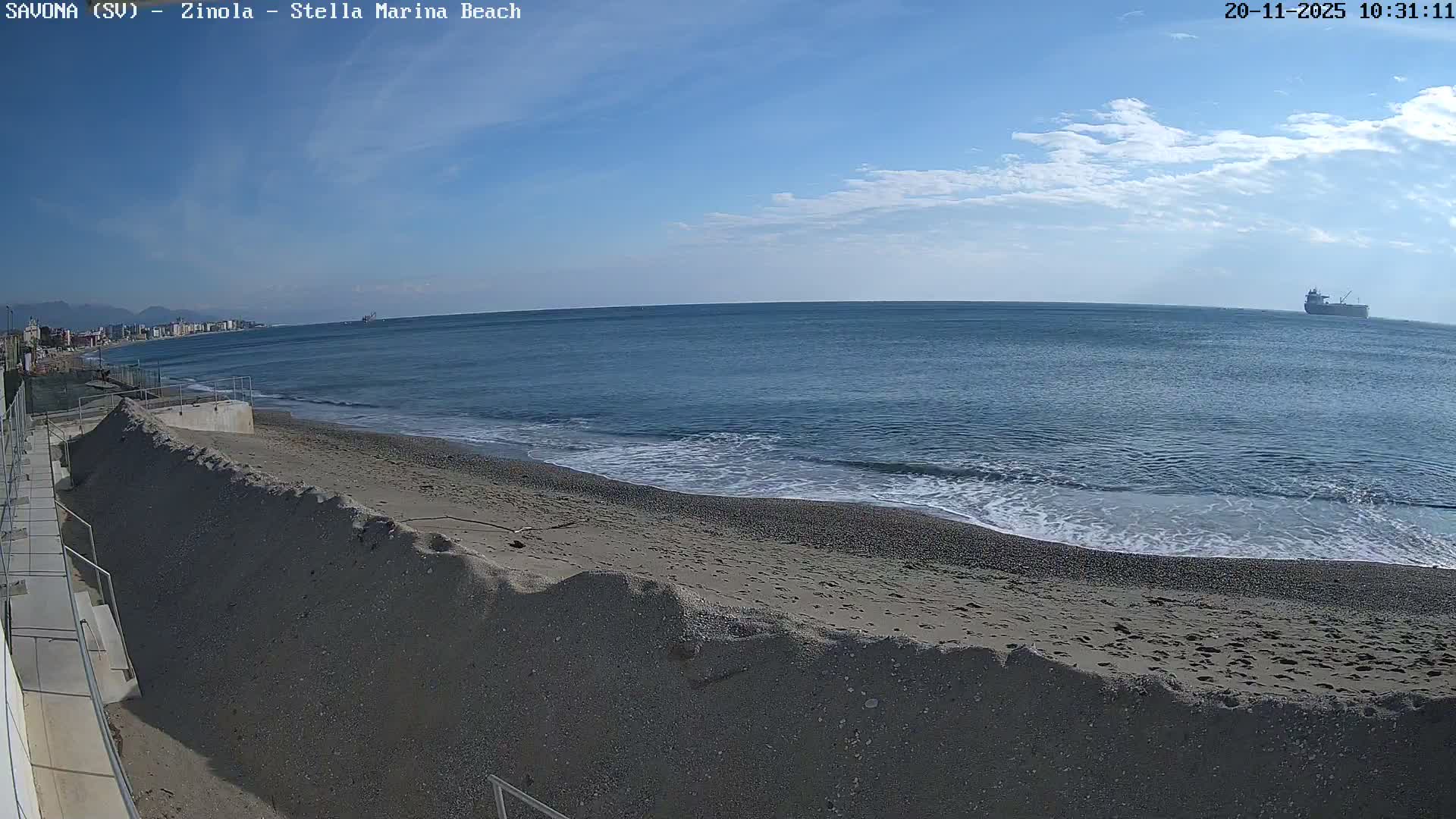 A clear, partly cloudy day illuminates a pebbly beach with gentle waves, a coastal town visible along the shoreline to the left, and a large cargo ship on the distant horizon to the right.