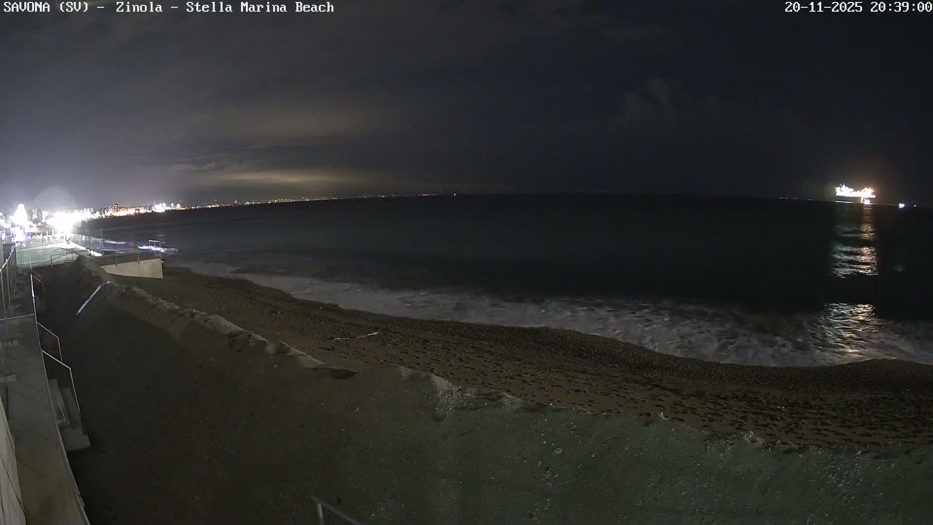 A cloudy night view captures a dark beach with gentle waves lapping the shore, a brightly lit coastal town in the distance to the left, and a large illuminated ship further out to sea on the right.