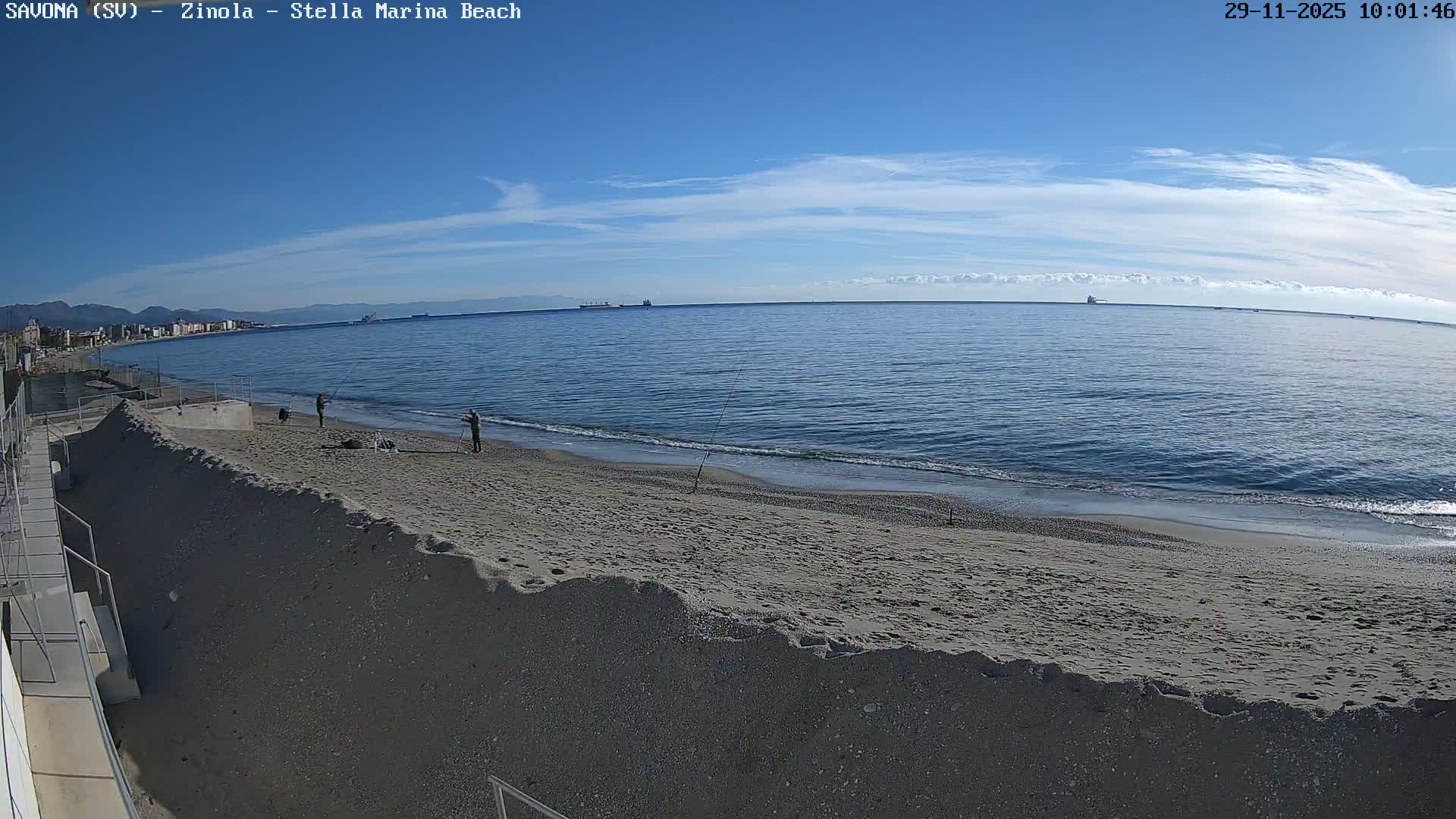 A wide view of a sunny beach shows two people fishing by the calm, blue sea, with a distant town and mountains along the coastline and a few ships on the horizon under a clear blue sky with sparse clouds.