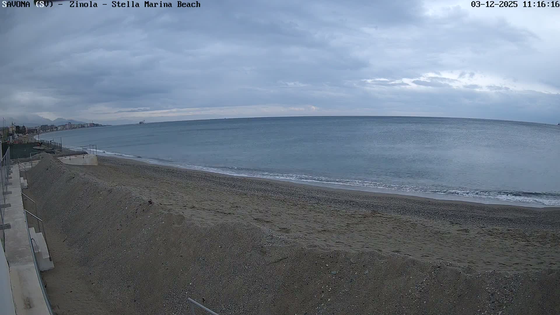 A wide view captures a calm, blue-grey sea gently lapping a broad greyish-brown sand and pebble beach, with a distant coastline of buildings and a vessel on the horizon, all under a heavily overcast sky.
