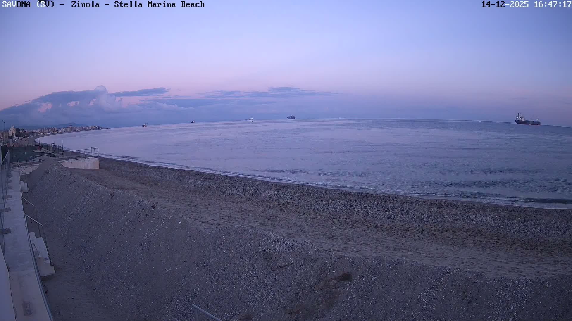 A wide view captures a calm, blue-grey sea gently lapping a broad greyish-brown sand and pebble beach, with a distant coastline of buildings and a vessel on the horizon, all under a heavily overcast sky.