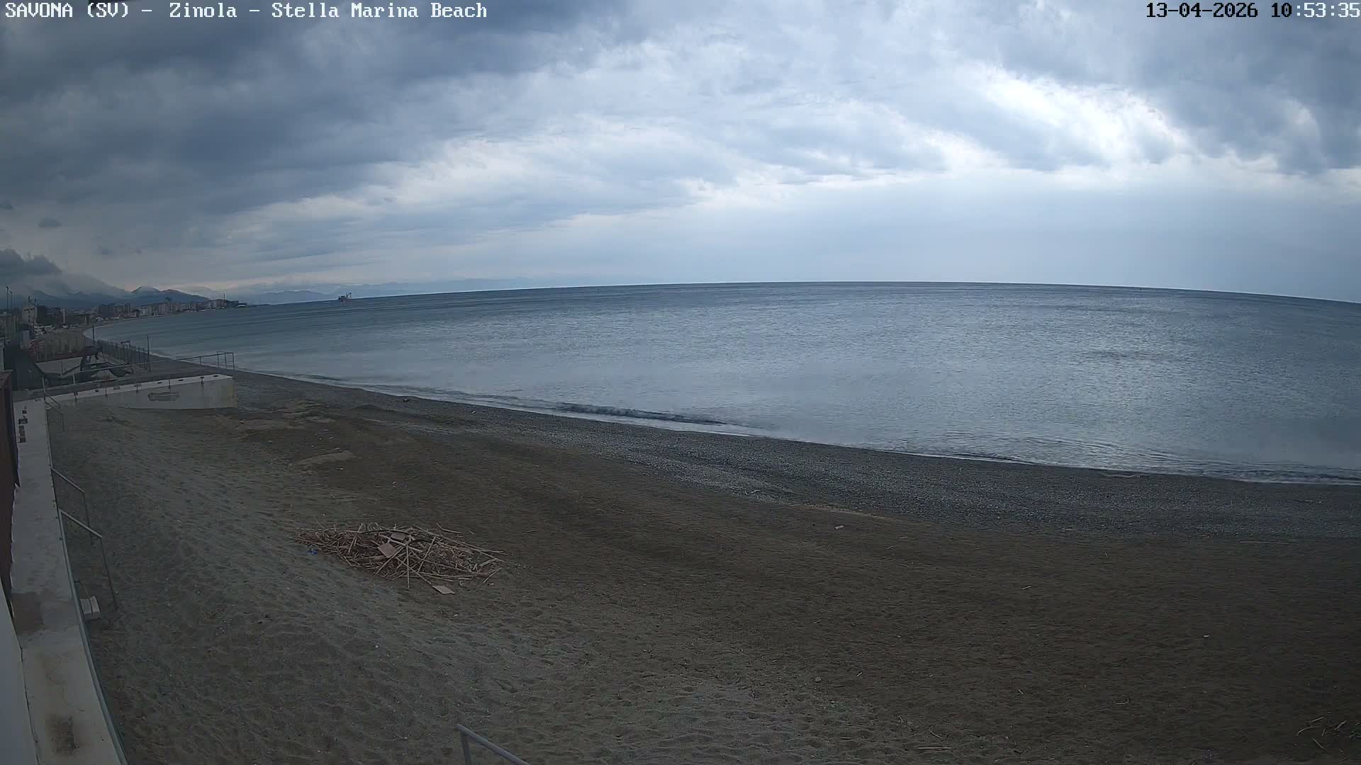 A wide view captures a calm, blue-grey sea gently lapping a broad greyish-brown sand and pebble beach, with a distant coastline of buildings and a vessel on the horizon, all under a heavily overcast sky.