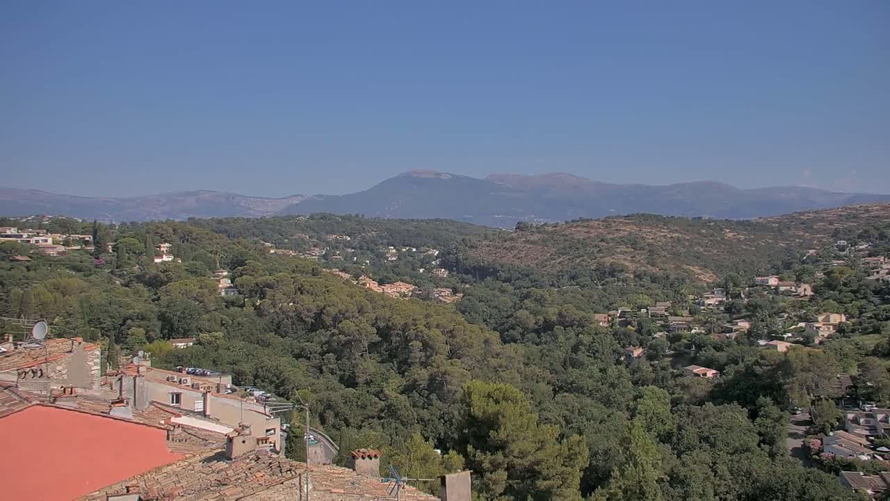 A sun-drenched hillside village nestled among lush green trees and rolling hills, with distant mountains under a clear blue sky.