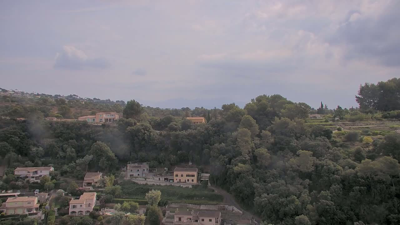 A hilly landscape with numerous houses nestled among lush green trees under a cloudy sky.