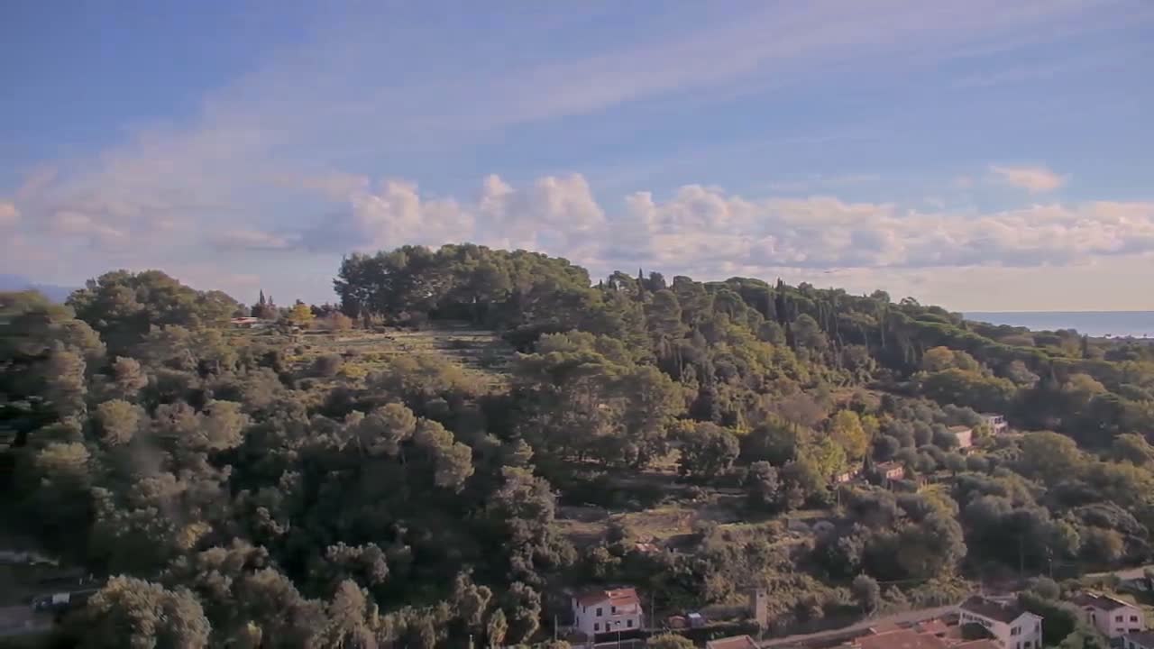 A sprawling, tree-covered hillside dotted with houses slopes down towards a distant sea under a partly cloudy blue sky.