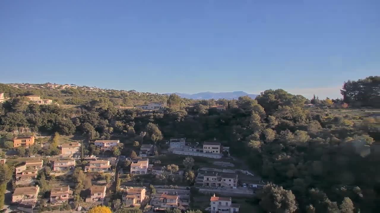 The image depicts a sunny day with clear blue skies over a hillside residential area densely covered with trees, with distant mountains visible on the horizon.