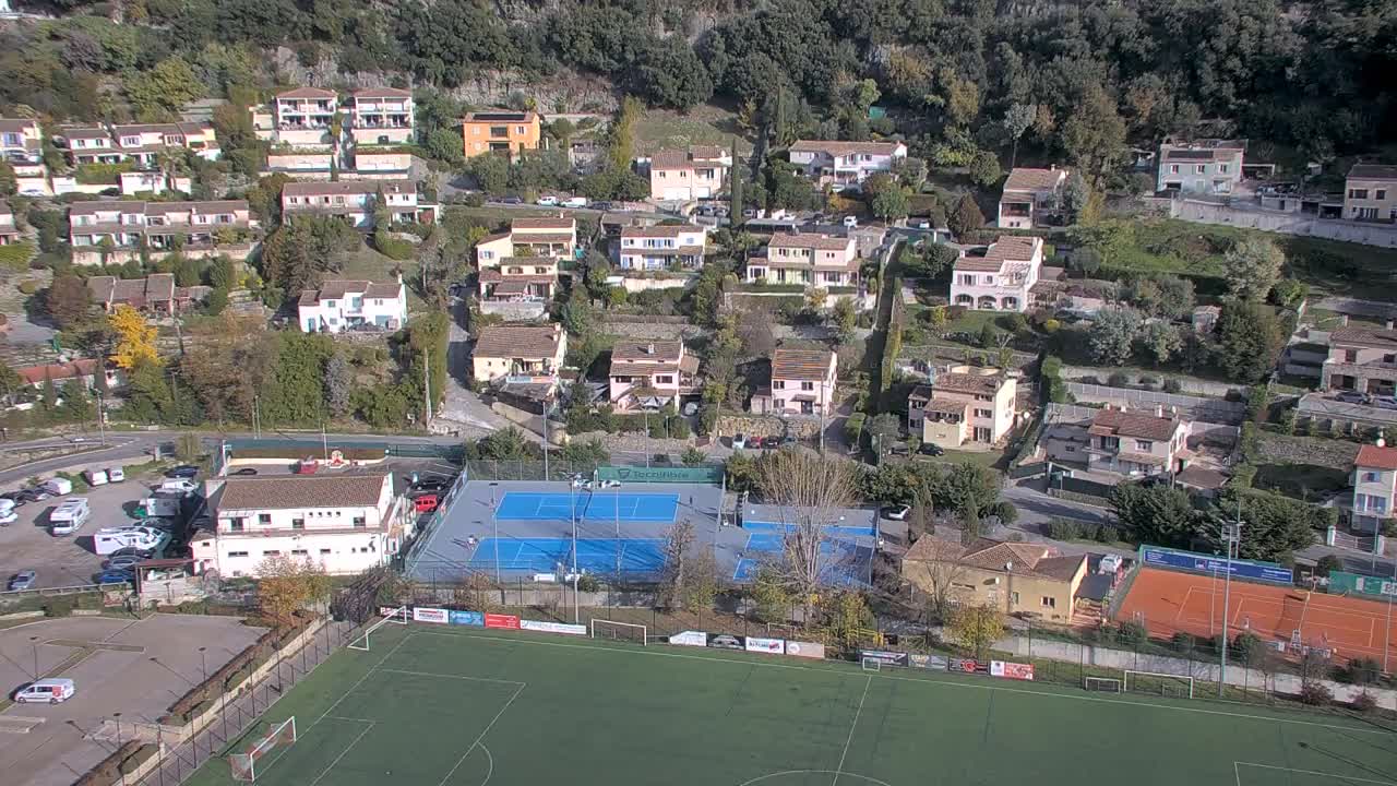 An aerial shot captures a sun-drenched hillside village featuring numerous terraced houses, a prominent green soccer field, and multiple blue and red tennis courts, all set against a backdrop of dense trees.