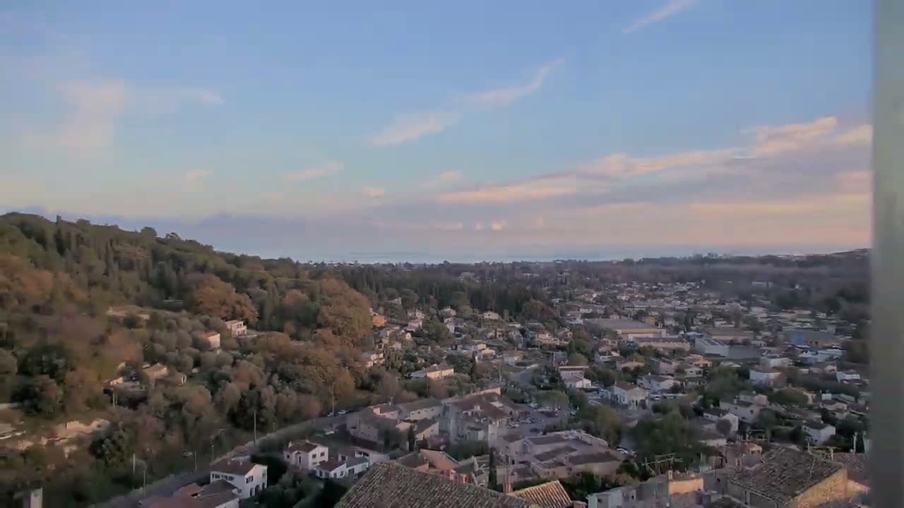 An aerial shot captures a sun-drenched hillside village featuring numerous terraced houses, a prominent green soccer field, and multiple blue and red tennis courts, all set against a backdrop of dense trees.