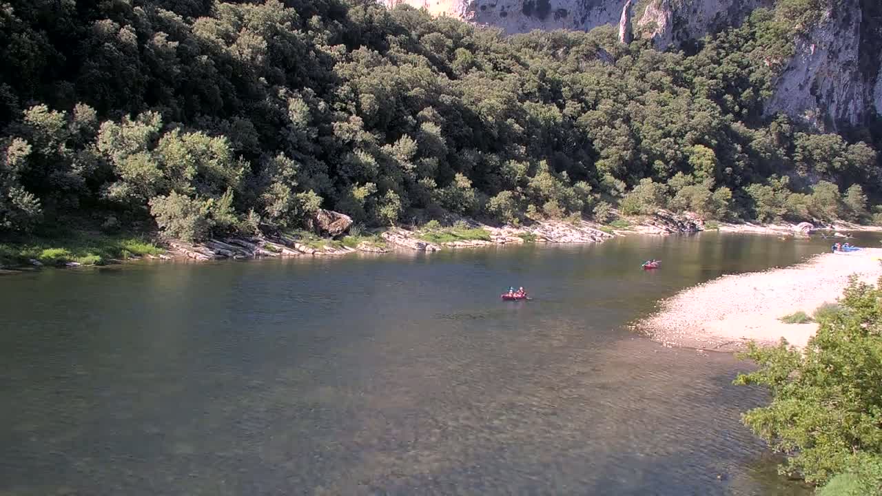 A calm river flows through a rocky, wooded valley under a sunny sky, with several small boats visible on the water.