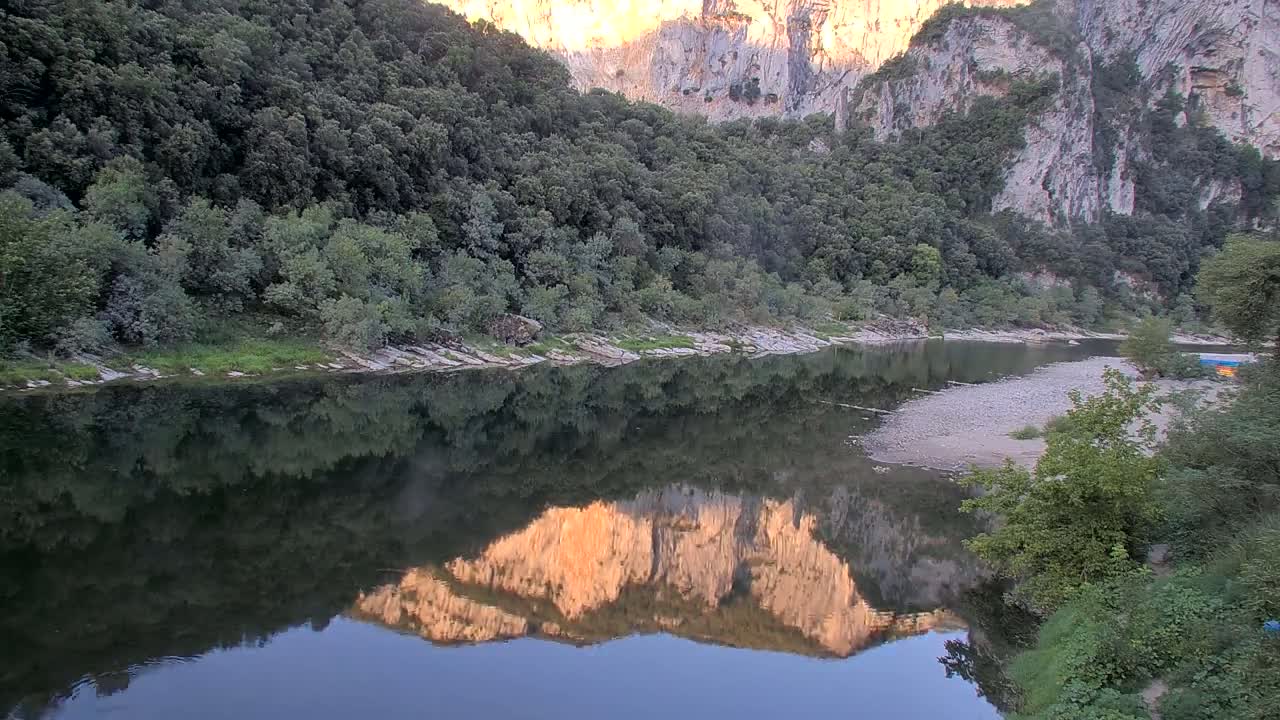 A calm river flows through a rocky, wooded valley under a sunny sky, with several small boats visible on the water.